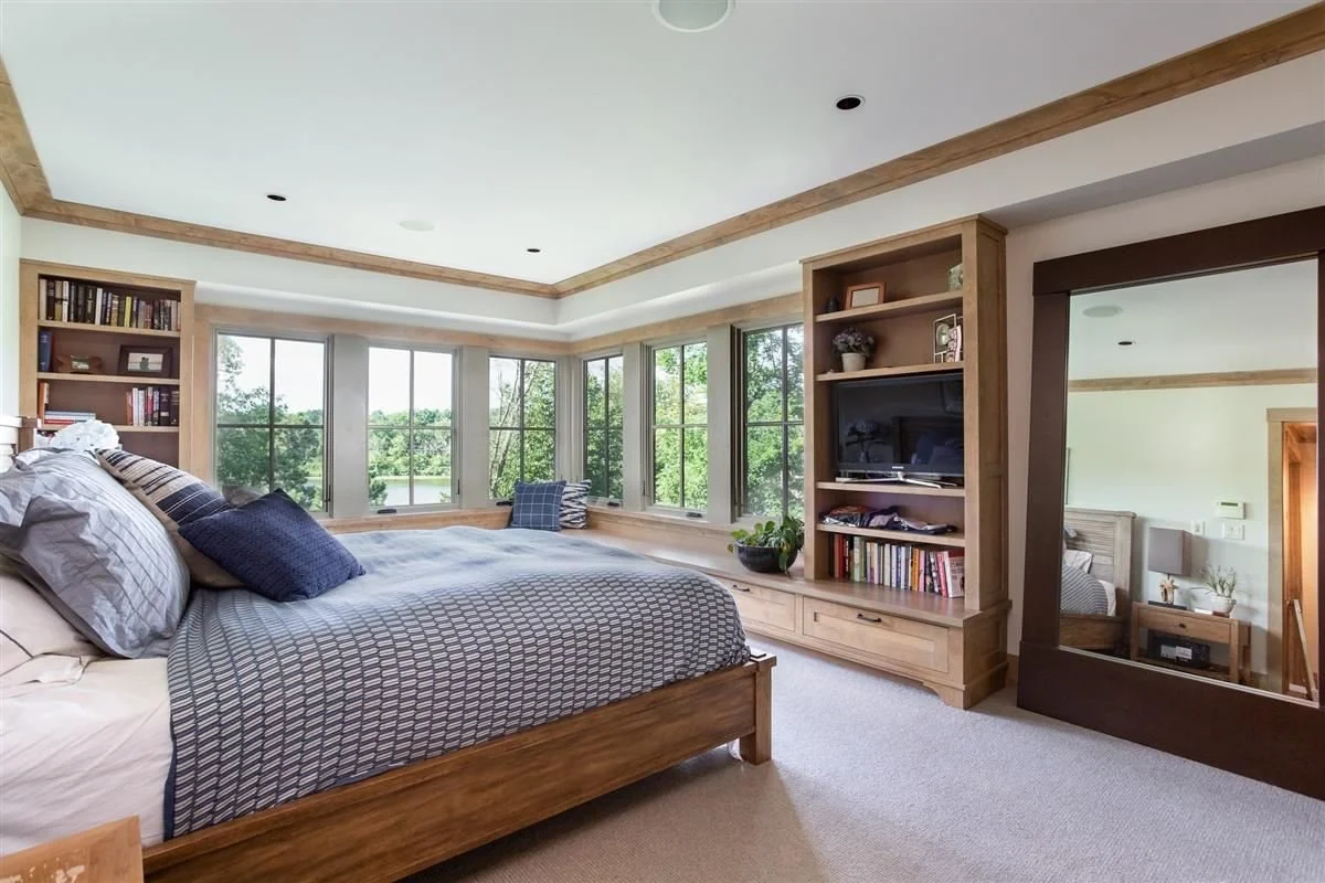 Bright bedroom with large windows overlooking trees, wooden furniture, bookshelves, a bed with pillows, and a large mirror.