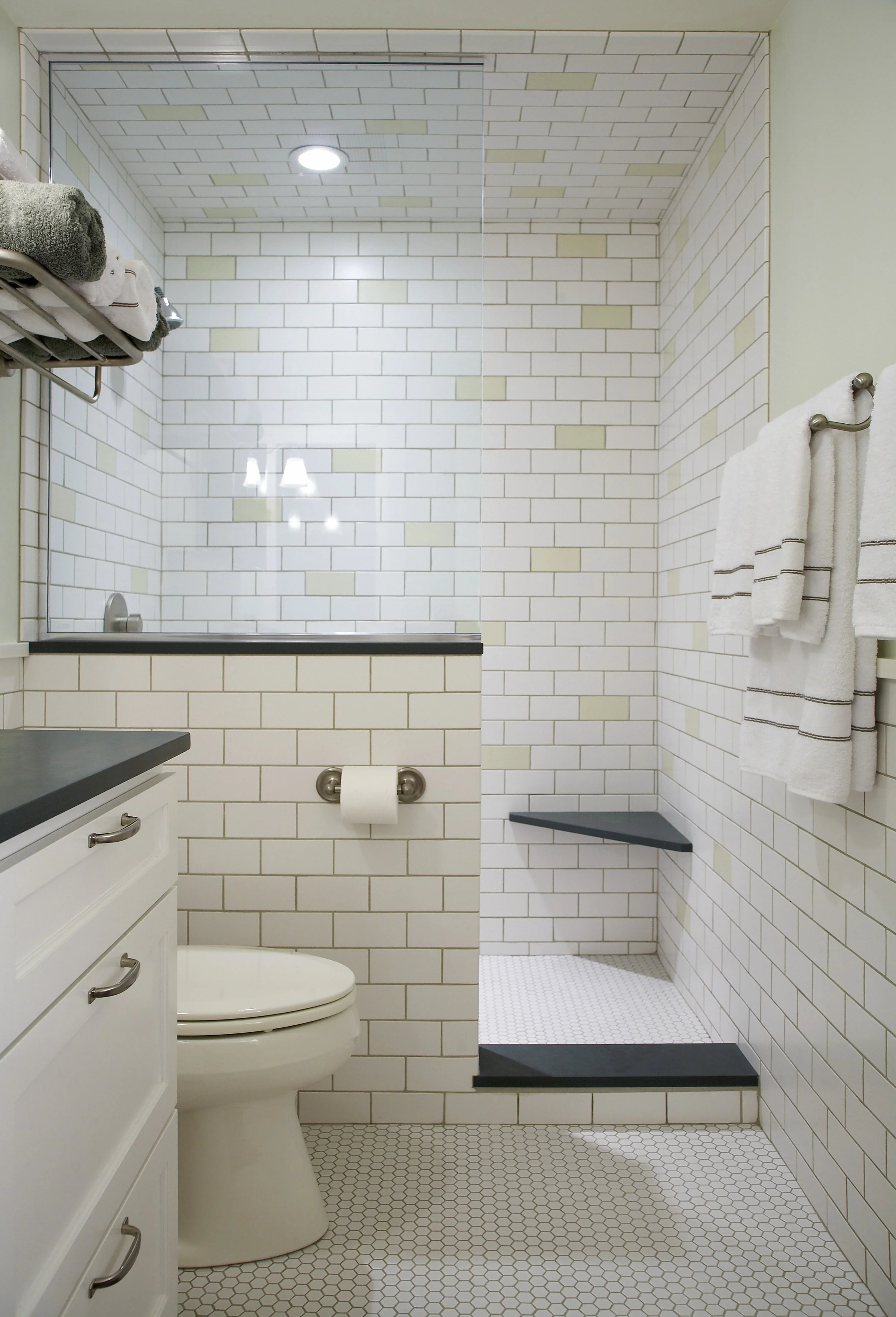 A bathroom with white subway tiles on the walls, a glass shower enclosure with a built-in shelf, a white toilet, a white vanity with drawers, a gray countertop, and towels hanging on a metal rod.
