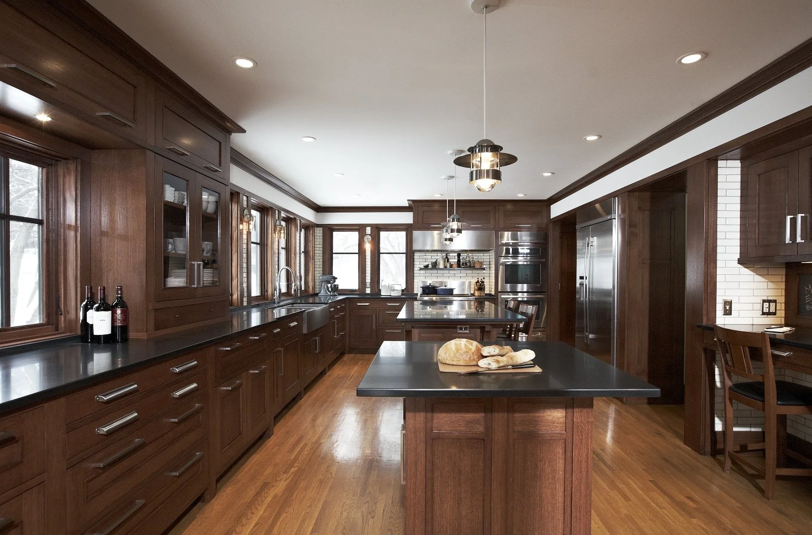 A spacious kitchen with dark wooden cabinets and black countertops. Multiple windows line the back wall, letting in natural light. There is an island with bread on a cutting board, and modern pendant lights hanging from the ceiling.