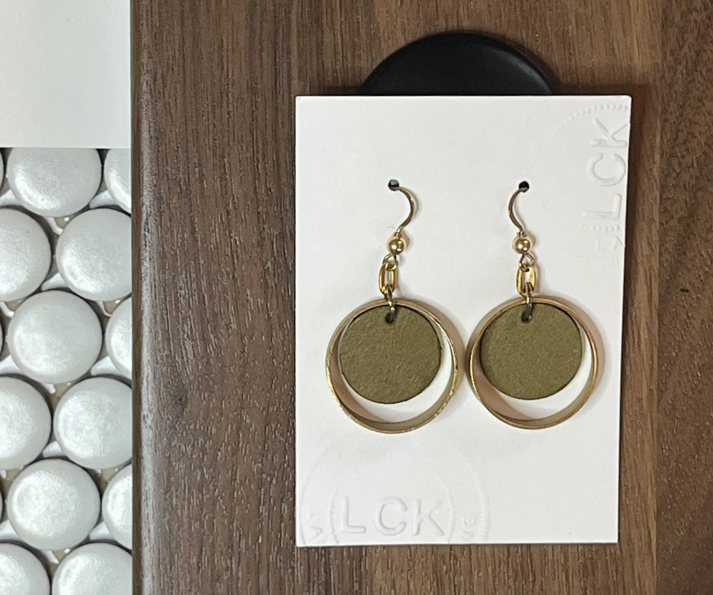 Close-up of a pair of gold and green earrings on a white card, placed on a wooden surface next to a white box with multiple round compartments.