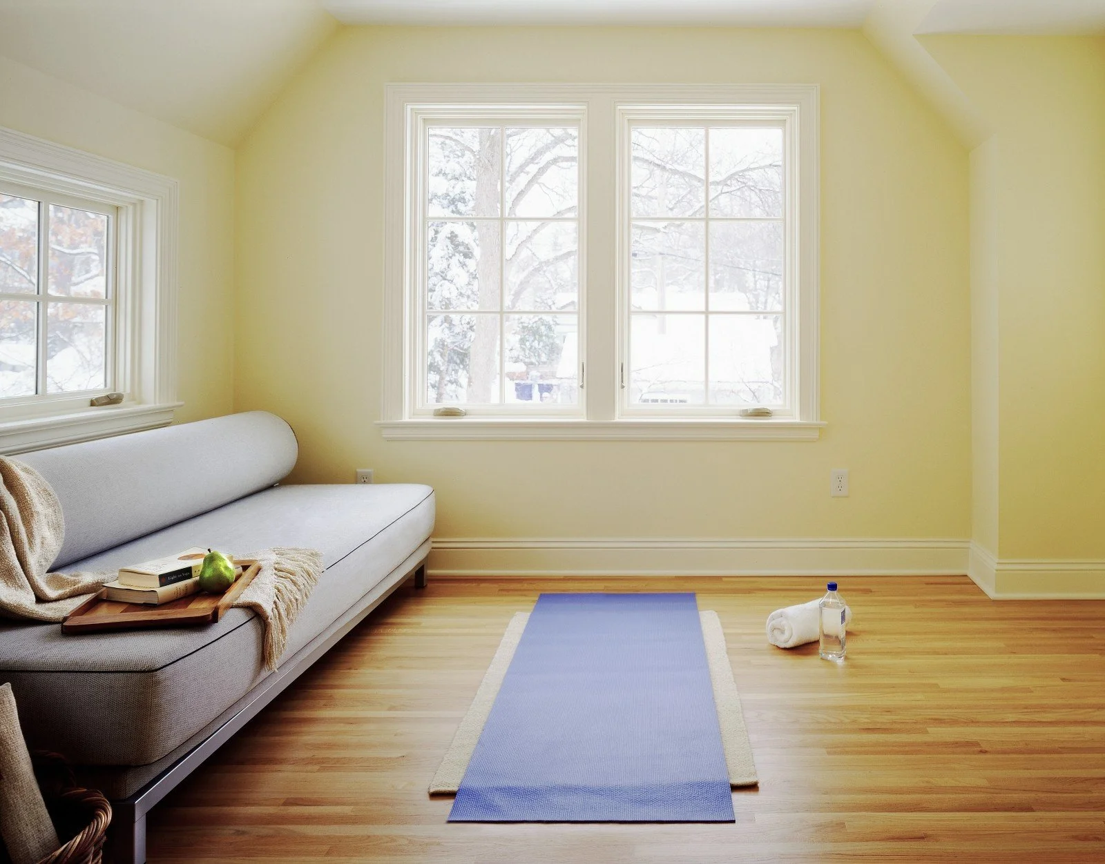A cozy room with yellow walls and wood flooring, featuring a large window with snowy trees outside. The room has a white sofa with a throw blanket and books on it, a yoga mat on the floor, and items like a rolled towel and a water bottle nearby.