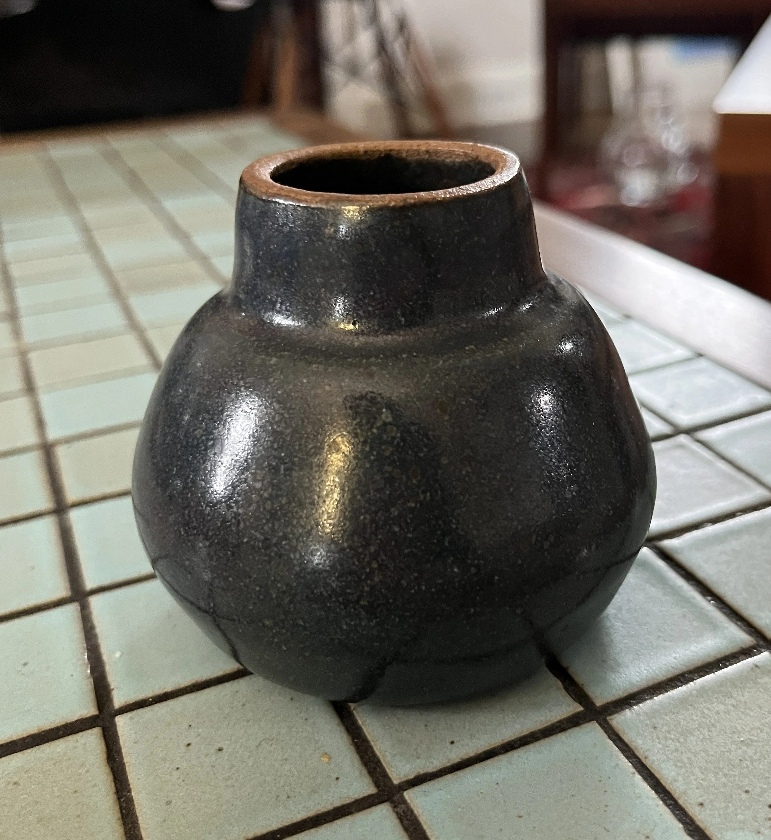 A black pottery vase with a wide opening and a textured surface, sitting on a tiled surface with white and brown grout lines.