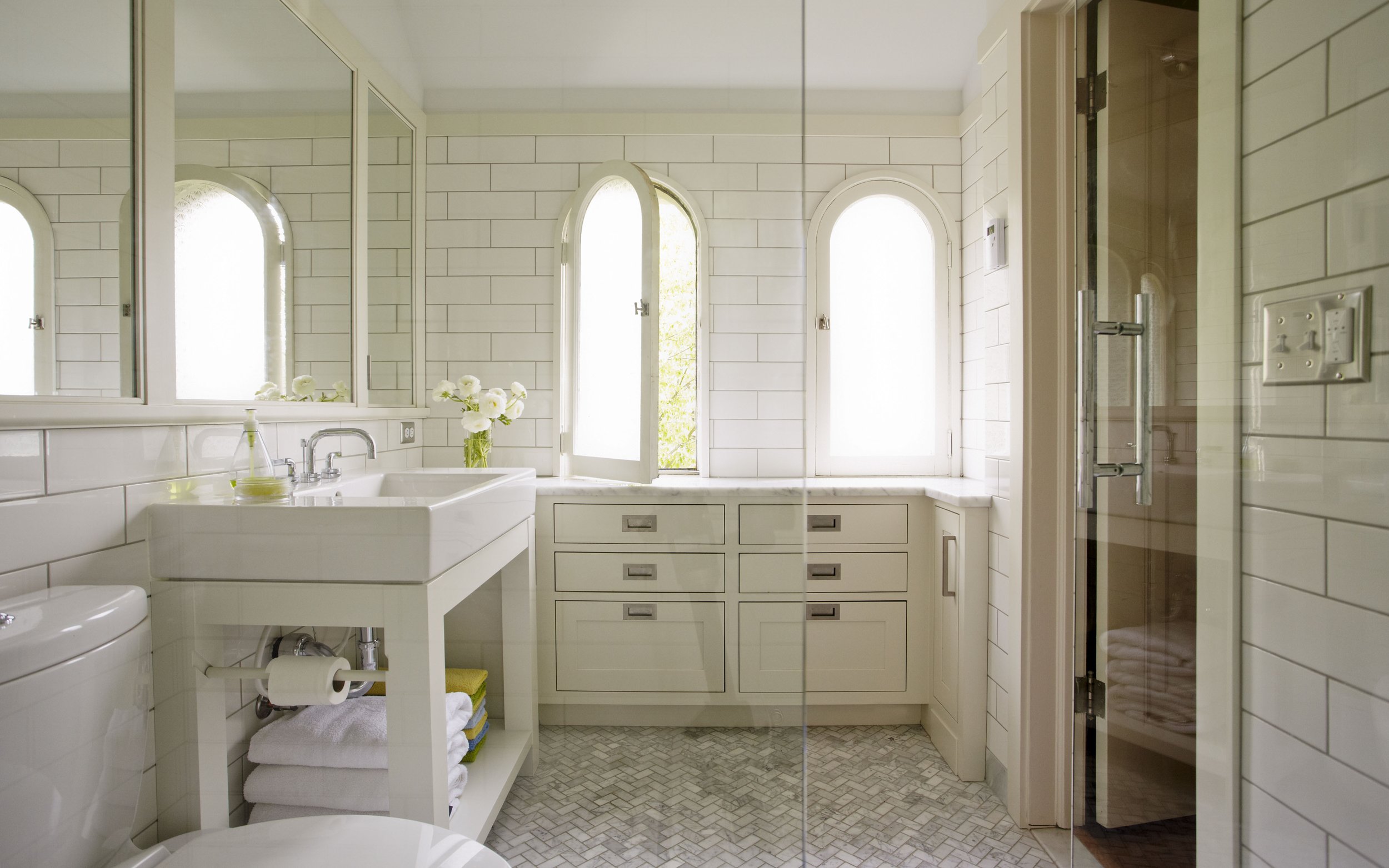 Bright bathroom with white subway tile walls, arched windows, and a white vanity with a small sink, flowers, towels, and a mirror.