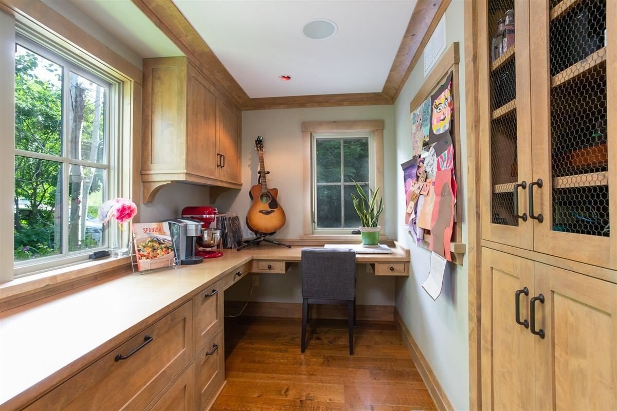 Home office with wooden cabinetry and floors, large windows with a view of greenery, a guitar hanging on the wall, a potted plant on the desk, and colorful paper crafts pinned on a corkboard.