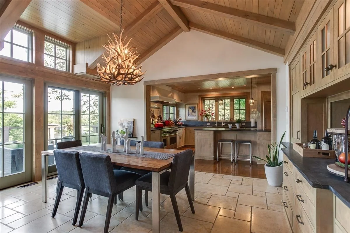 A dining area with a wooden table and six dark upholstered chairs, a deer antler chandelier hanging from a wooden ceiling, next to large windows and a door leading outside. The kitchen in the background has wooden cabinetry, a countertop, and a windo