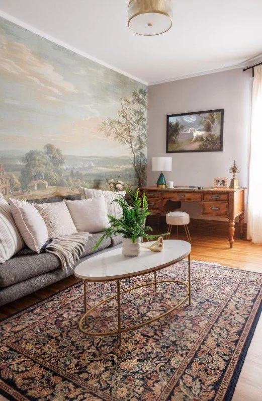 Living room with a gray sofa, a white oval coffee table with a potted plant, a vintage-style rug, a wooden sideboard, a lamp, a framed landscape painting, and a large wall mural depicting an outdoor scene with trees and a sky.