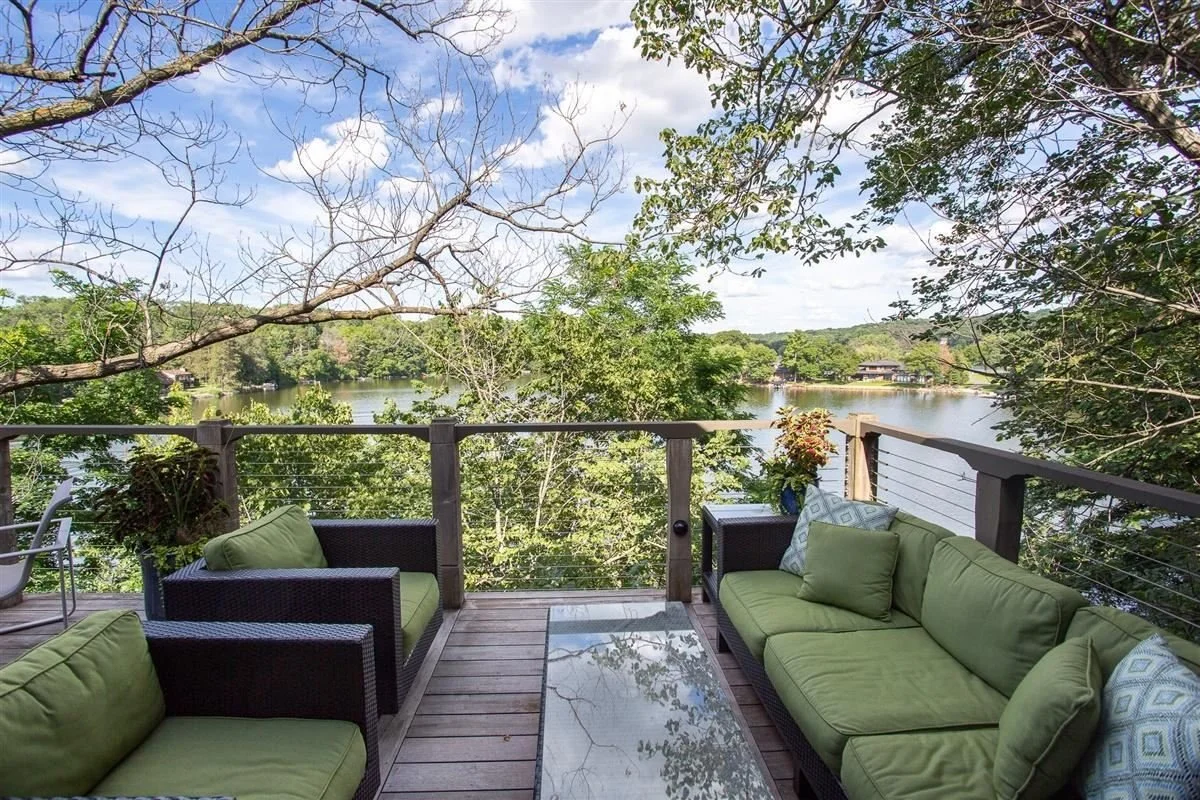 View of a lakeside porch with green cushioned seating and a glass table, surrounded by trees and overlooking a lake with houses on the opposite shore.