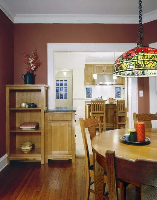 Dining area with wooden table and chairs, colorful stained-glass pendant light, and a bookshelf with decorative items and books, leading to a kitchen with wooden cabinets and bar stools, in a home interior.