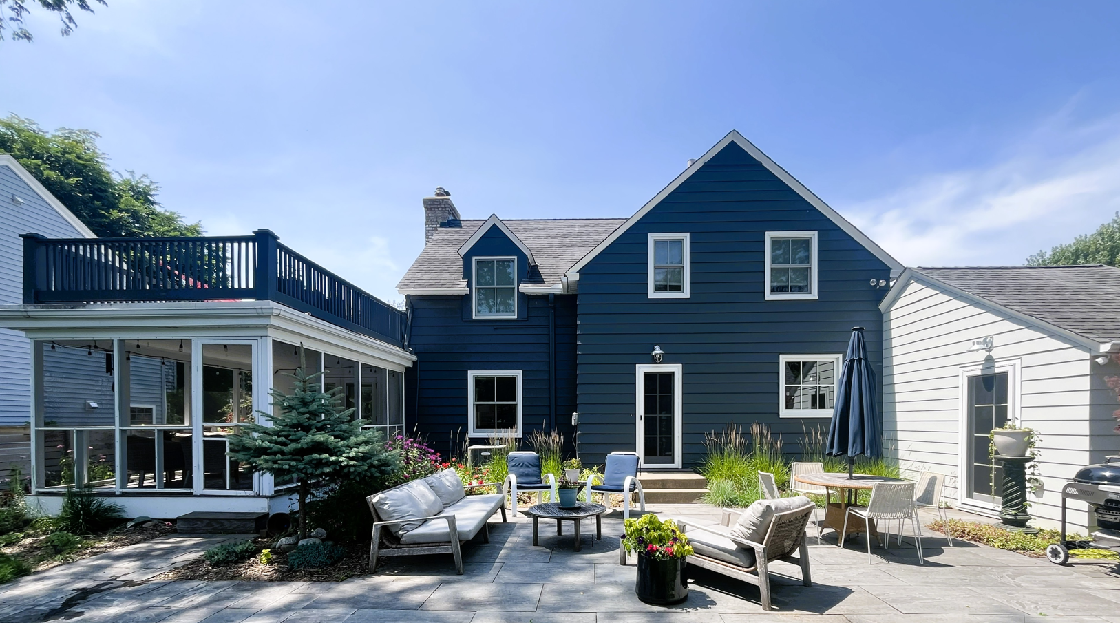 Backyard patio with outdoor furniture, a small pine tree, garden plants, and a blue house with multiple stories and a deck.