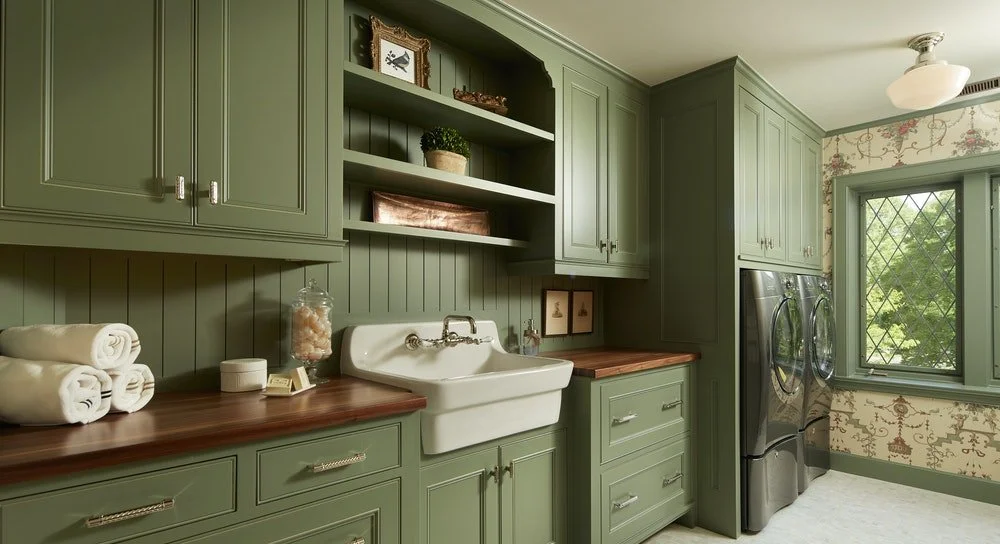Green laundry room with a white farmhouse sink, front-loading washer and dryer, open shelves with decor, window with view outside, patterned wallpaper, and wooden countertops.