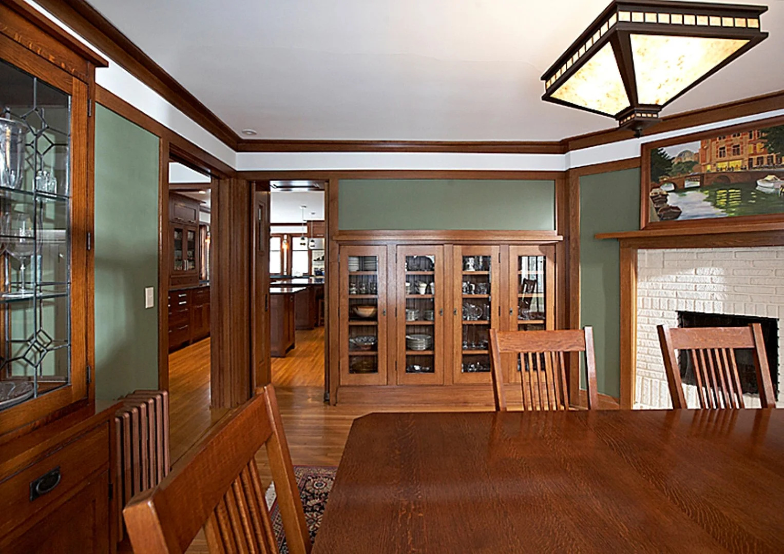 A dining room with a wooden table and chairs, a built-in china cabinet with glass doors, a brick fireplace, a large painting above the fireplace, and a ceiling light fixture. The walls are painted green with wooden trim.