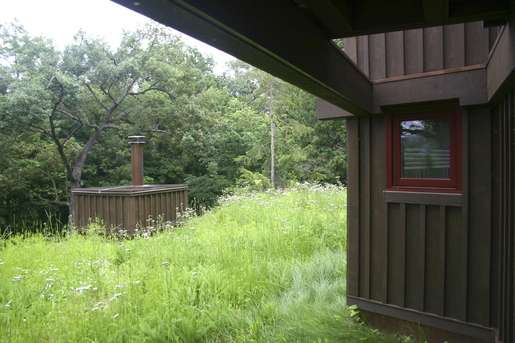 Side of a wooden house with a window, surrounded by green grass and wildflowers, with trees in the background