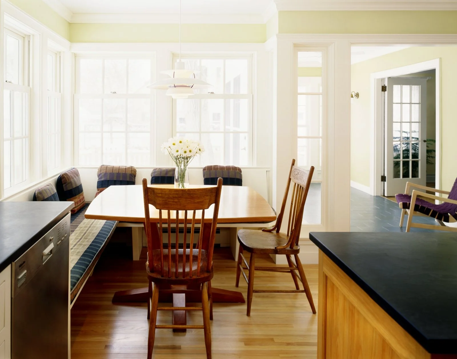 Bright dining area with large windows, a wooden table, mismatched chairs, a flower vase, and patio doors leading outside.