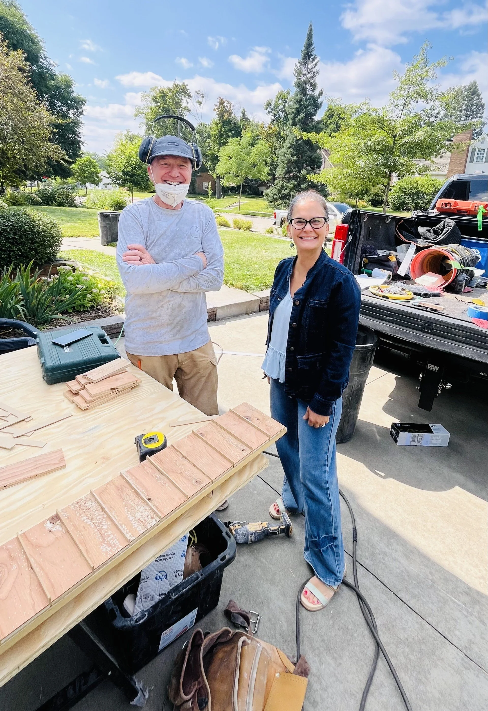 Two people outdoors, one man wearing a helmet with ear protection and a face mask, and a woman wearing glasses, standing near a work table with bricks and tools, in a sunny residential neighborhood with trees and parked cars.