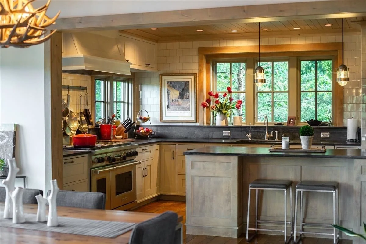 A cozy kitchen with cream-colored cabinets, black countertop, and a large window with a view of greenery. There are two hanging pendant lights above the counter, red flowers in a vase, and a potted plant. A framed picture is on the wall, and various 