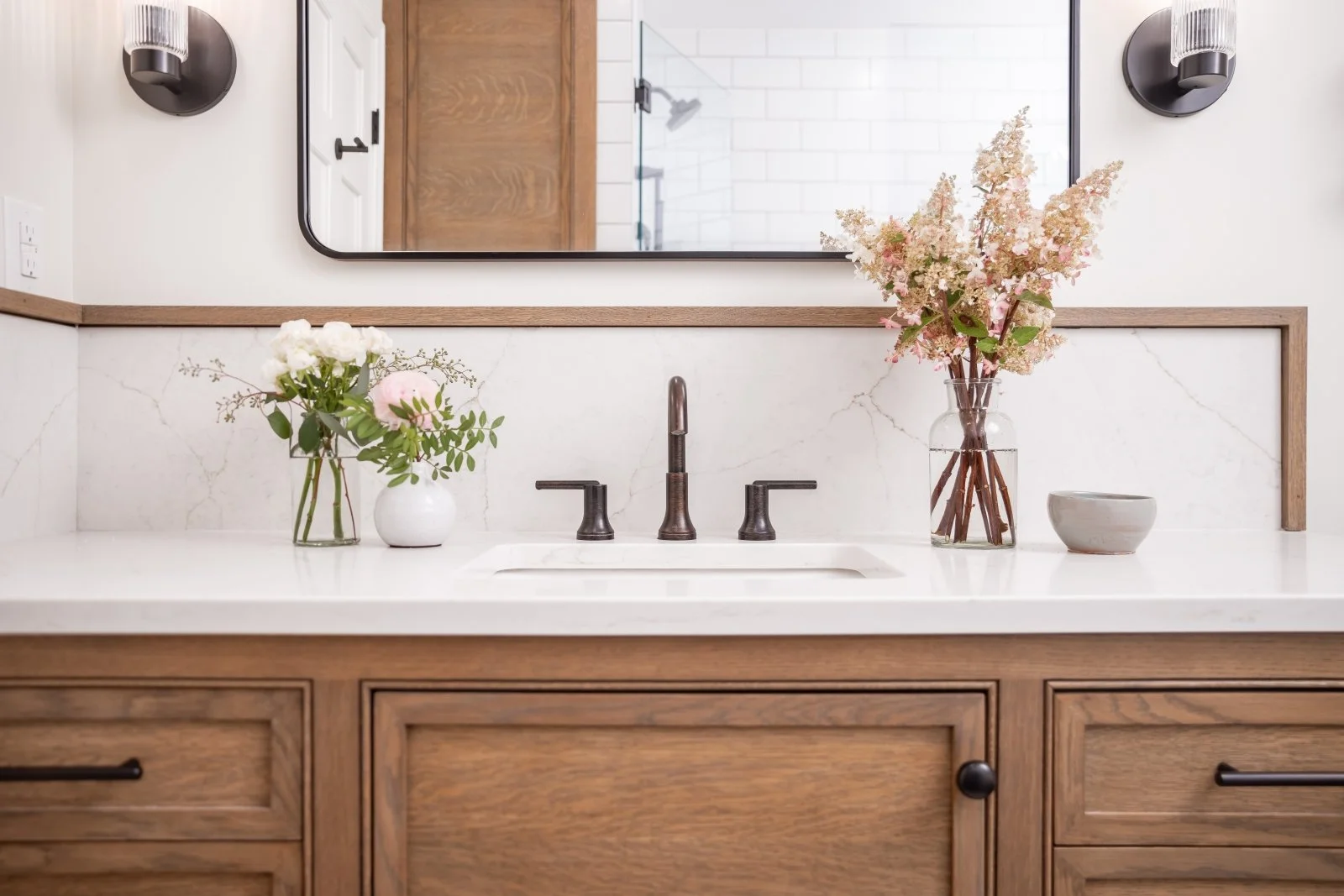 Bathroom vanity with a white countertop, two glass vases with pink and white flowers, a small ceramic bowl, a mirror, and black hardware, with a shower visible in the mirror reflection.