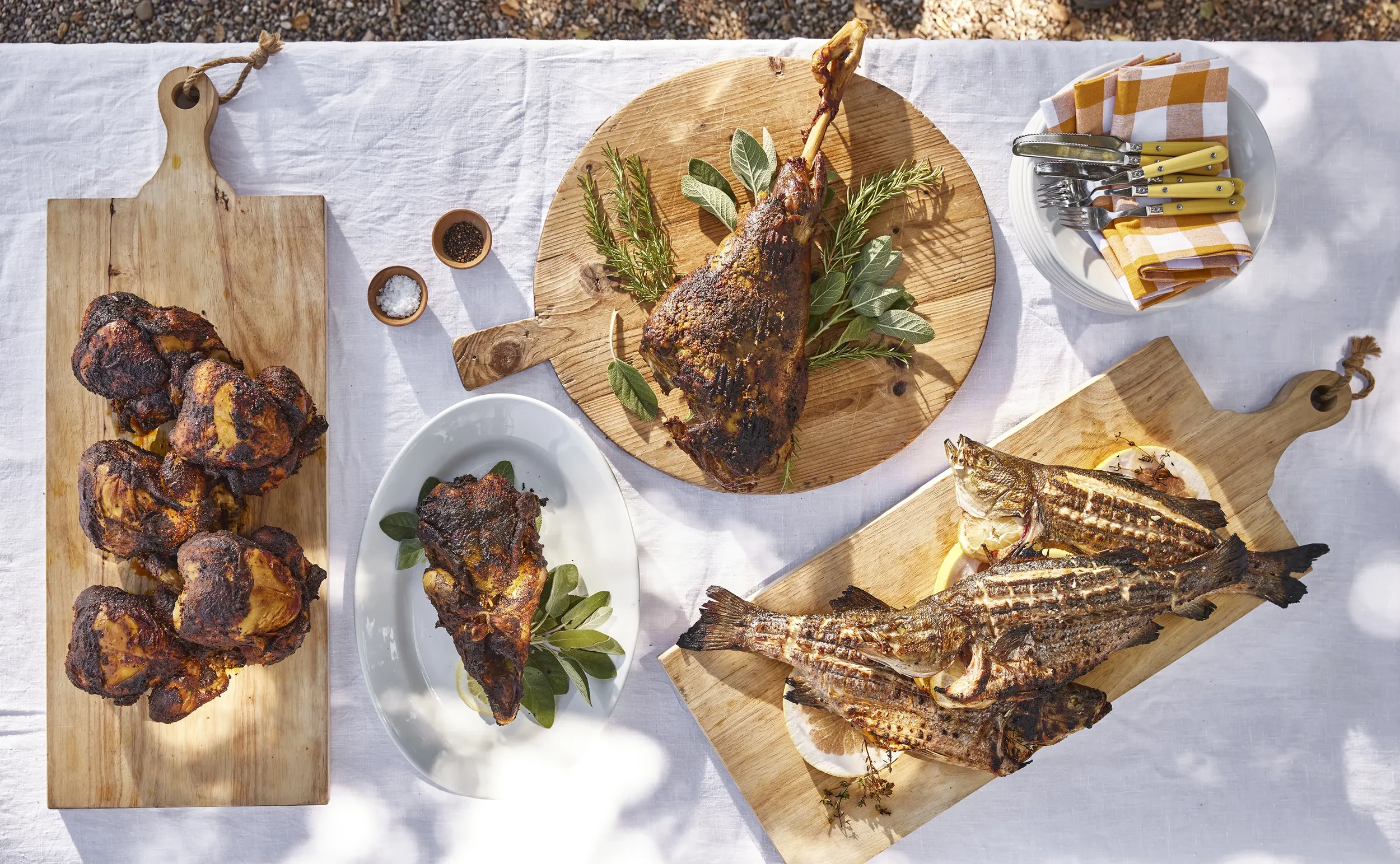 A spread of grilled meats on wooden boards, including chicken drumsticks, a lamb chop on a round wooden cutting board, and grilled whole fish on a rectangular wooden board, with plates of utensils, small bowls of spices, and a napkin with cutlery on 