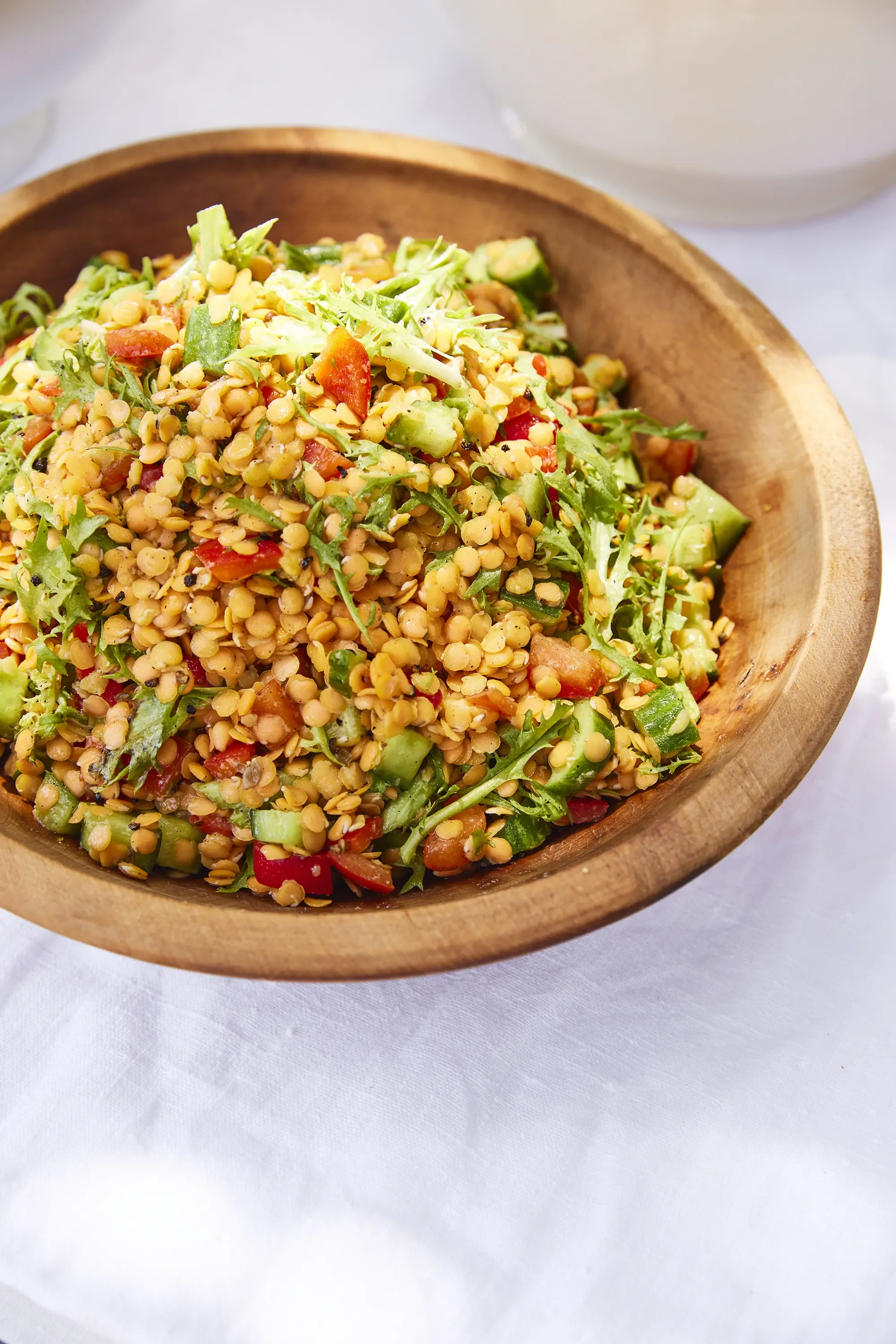 A wooden bowl filled with lentil salad containing chopped vegetables like tomatoes and cucumbers, placed on a white tablecloth.