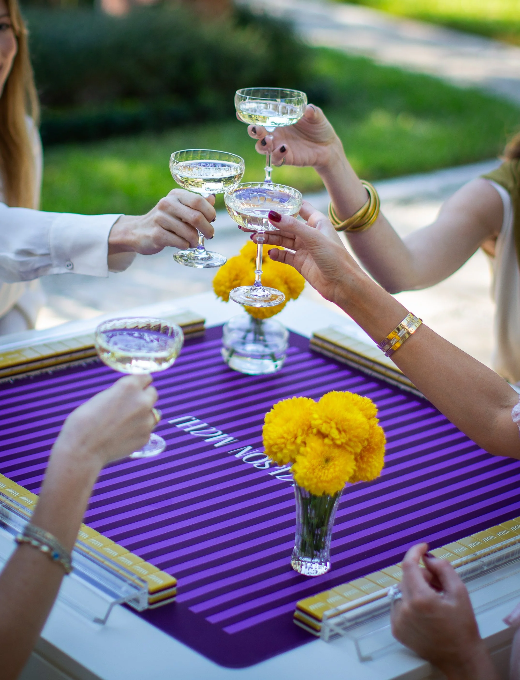 People clinking champagne glasses with a purple-striped tablecloth, yellow flowers in a clear vase, and yellow and white mitm tray at an outdoor gathering.