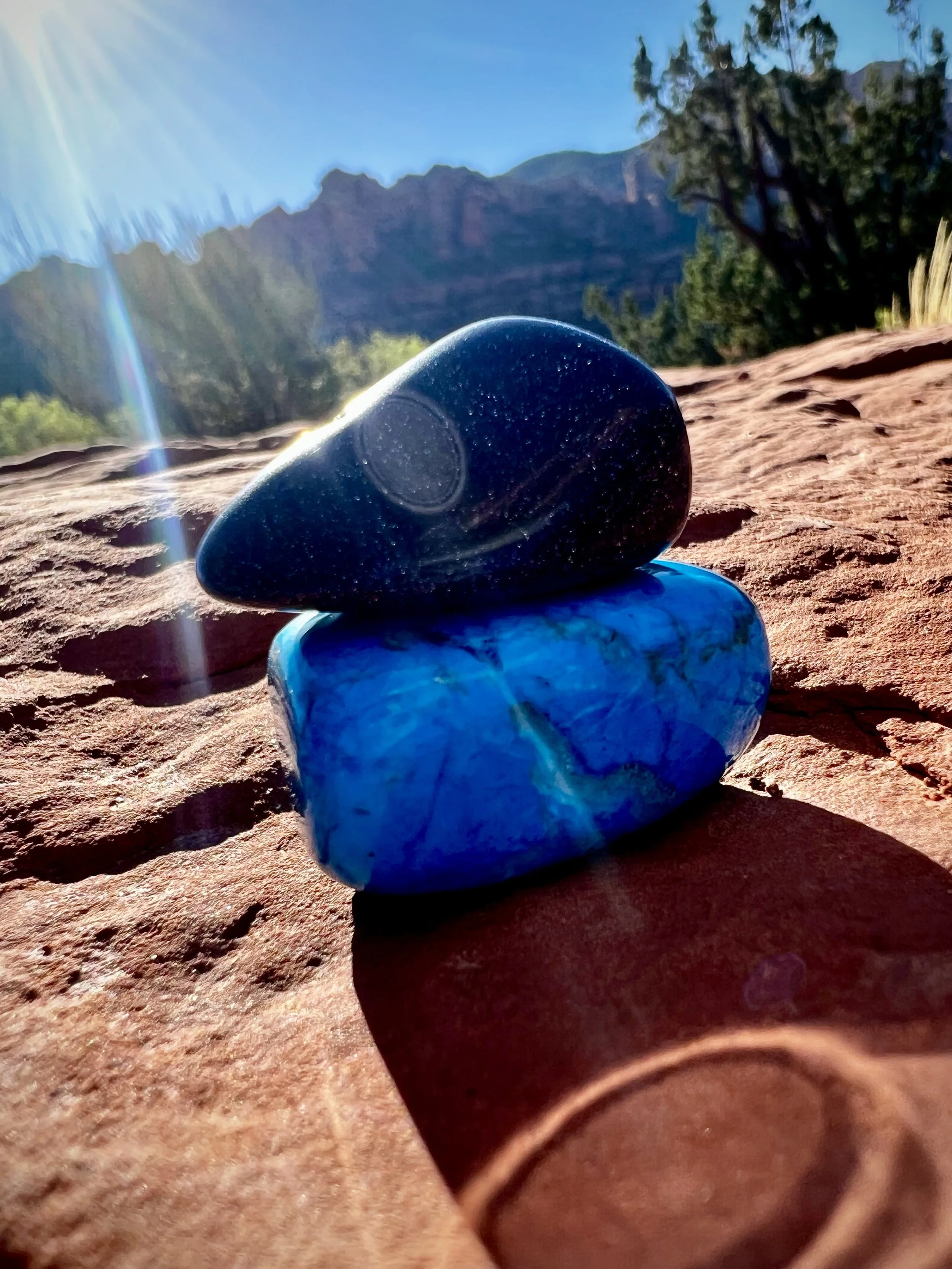 Close-up of two stacked stones, one black and one blue, on a reddish rock surface with a desert landscape, mountains, and a clear blue sky in the background, with sunlight creating lens flares.