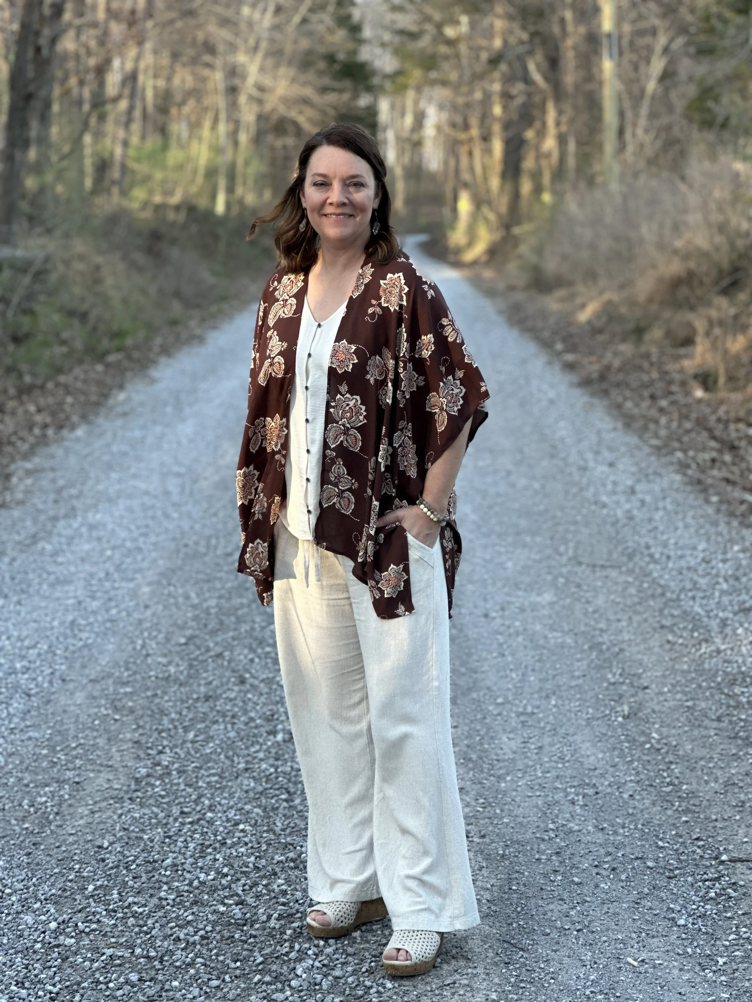 A woman standing on a gravel road in a forested area during daylight, smiling, wearing a floral patterned brown kimono, white top, cream wide-leg pants, and wedge sandals.