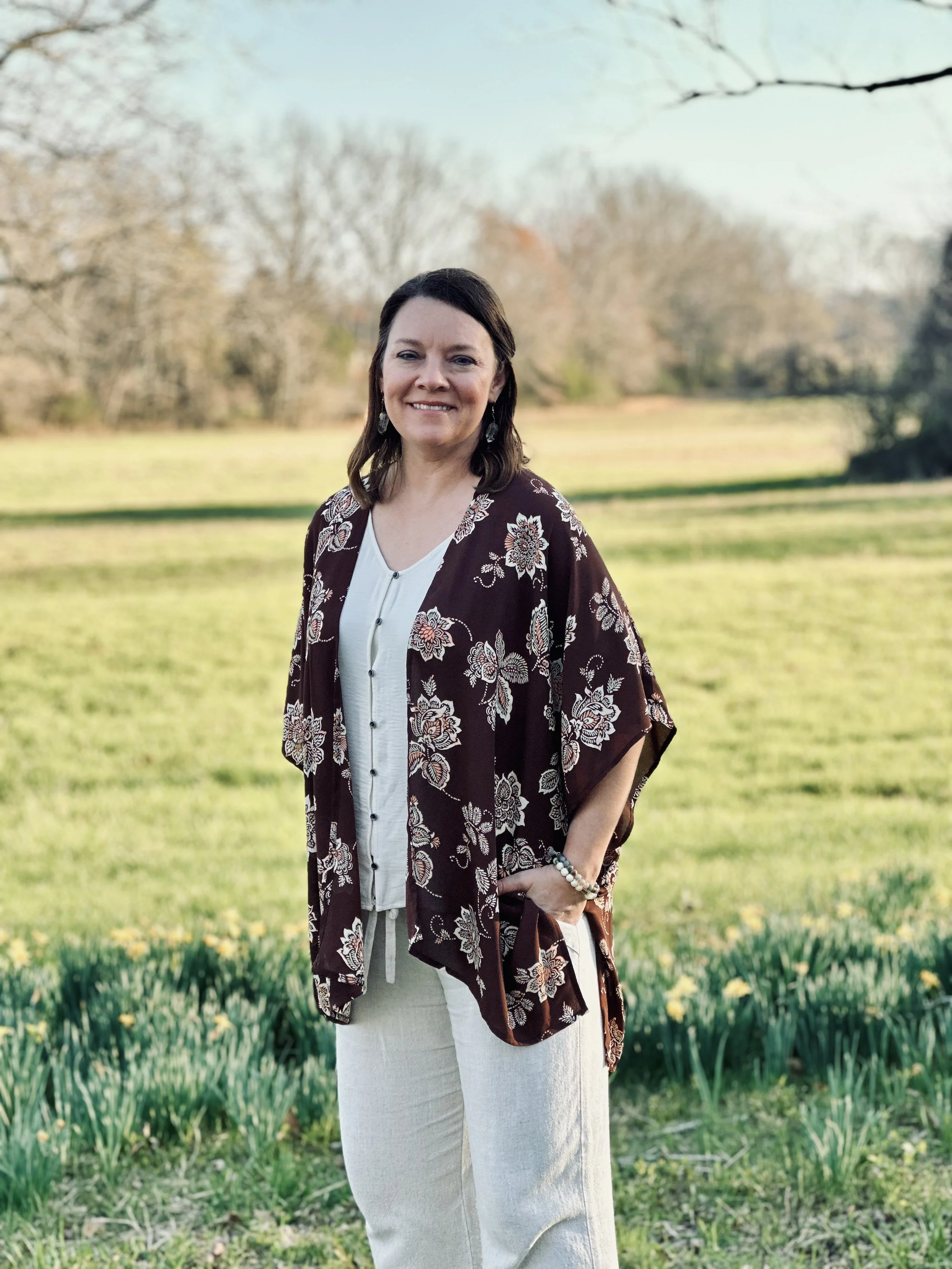 A woman standing outdoors on a grassy field during daytime, smiling at the camera, with trees and a clear sky in the background.