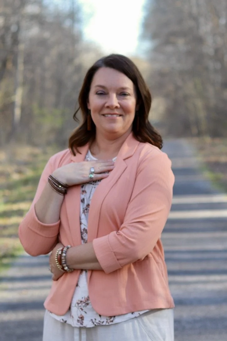 A woman standing on a tree-lined road, wearing a peach blazer and white patterned blouse, smiling with her right hand on her chest, and wearing jewelry including rings, bracelets, and earrings.