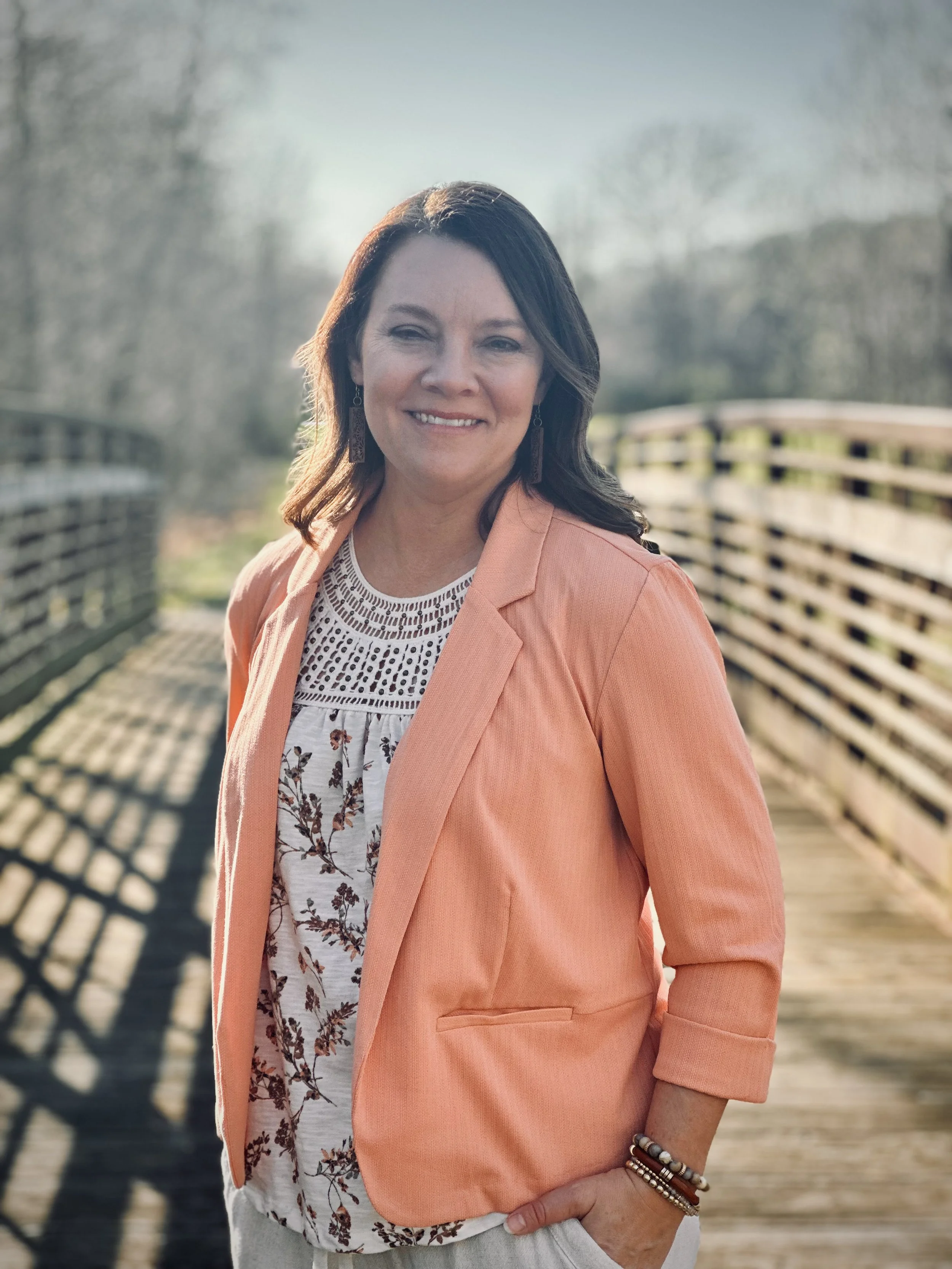A woman with shoulder-length dark hair smiling outdoors on a wooden bridge, wearing a peach blazer and patterned blouse with floral design.