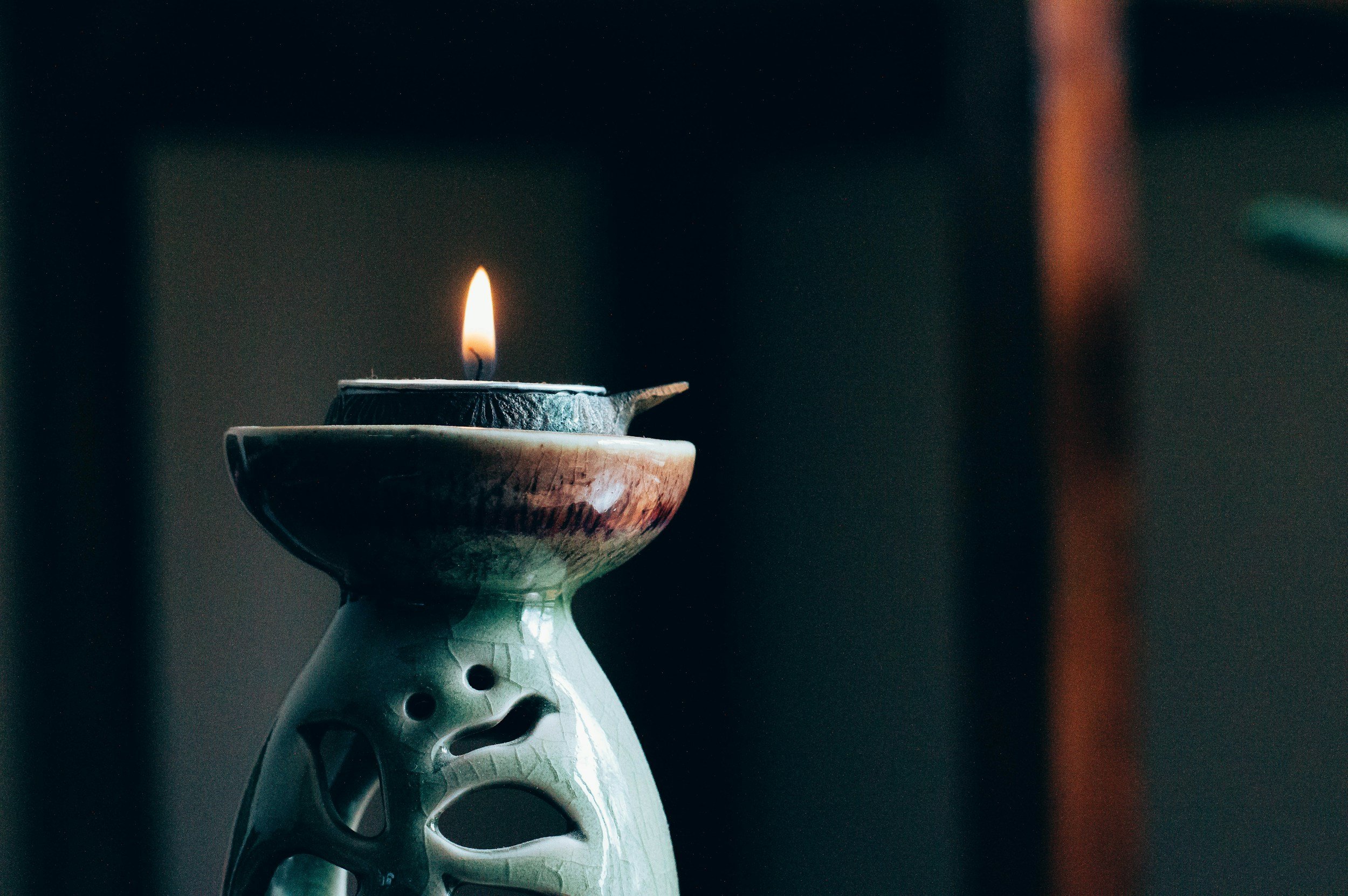 A lit candle in a ceramic holder with a face design, placed on a matching ceramic dish, against a dark blurred background.