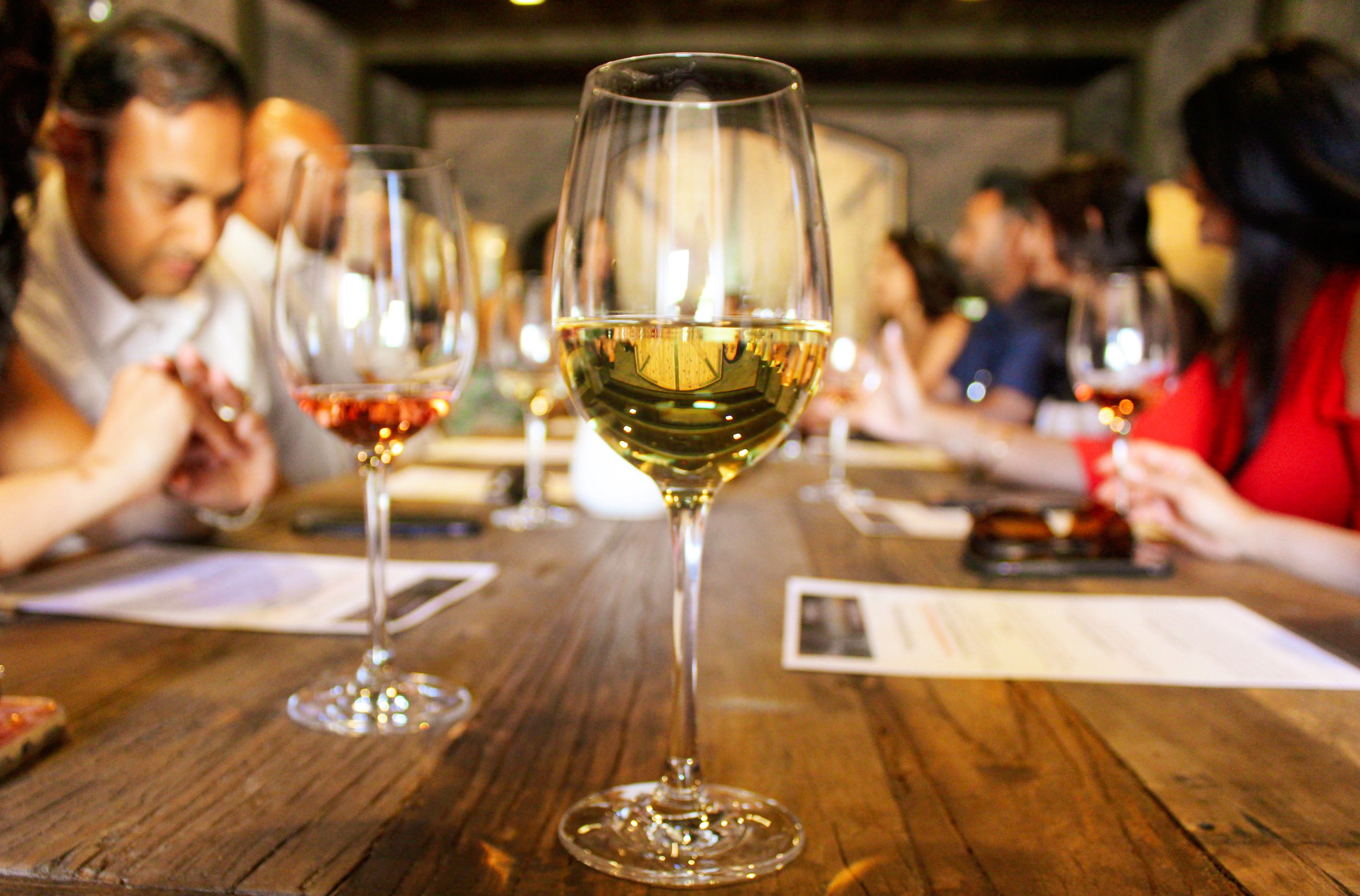 A close-up of a glass of white wine on a wooden table with people dining in the background at a winery.