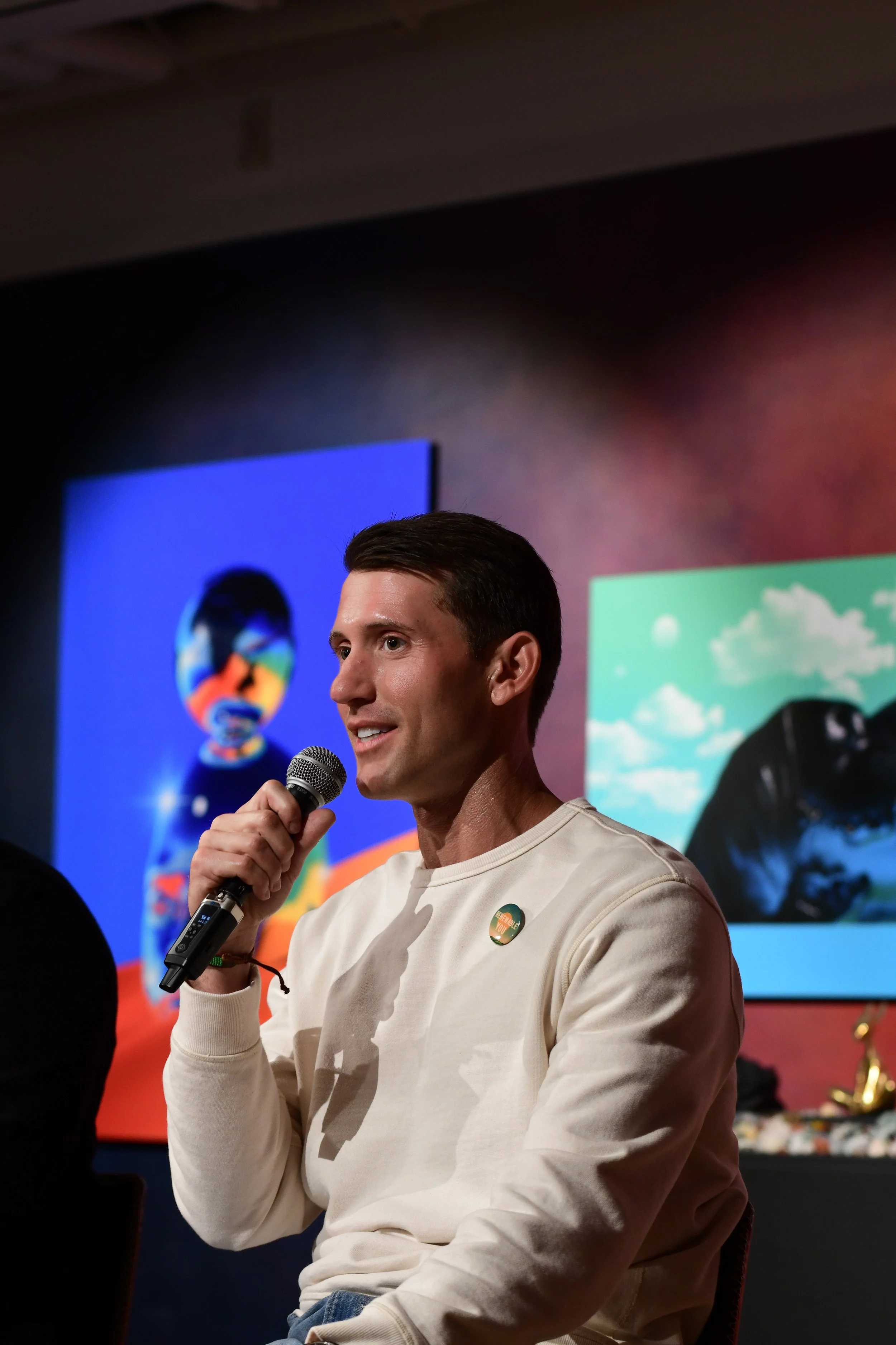 A man tech entreprenur with short dark hair wearing a beige sweatshirt and holding a microphone speaks at an event. Behind him, there is a colorful abstract images and a rainbow-colored push pin badge on his sweatshirt.