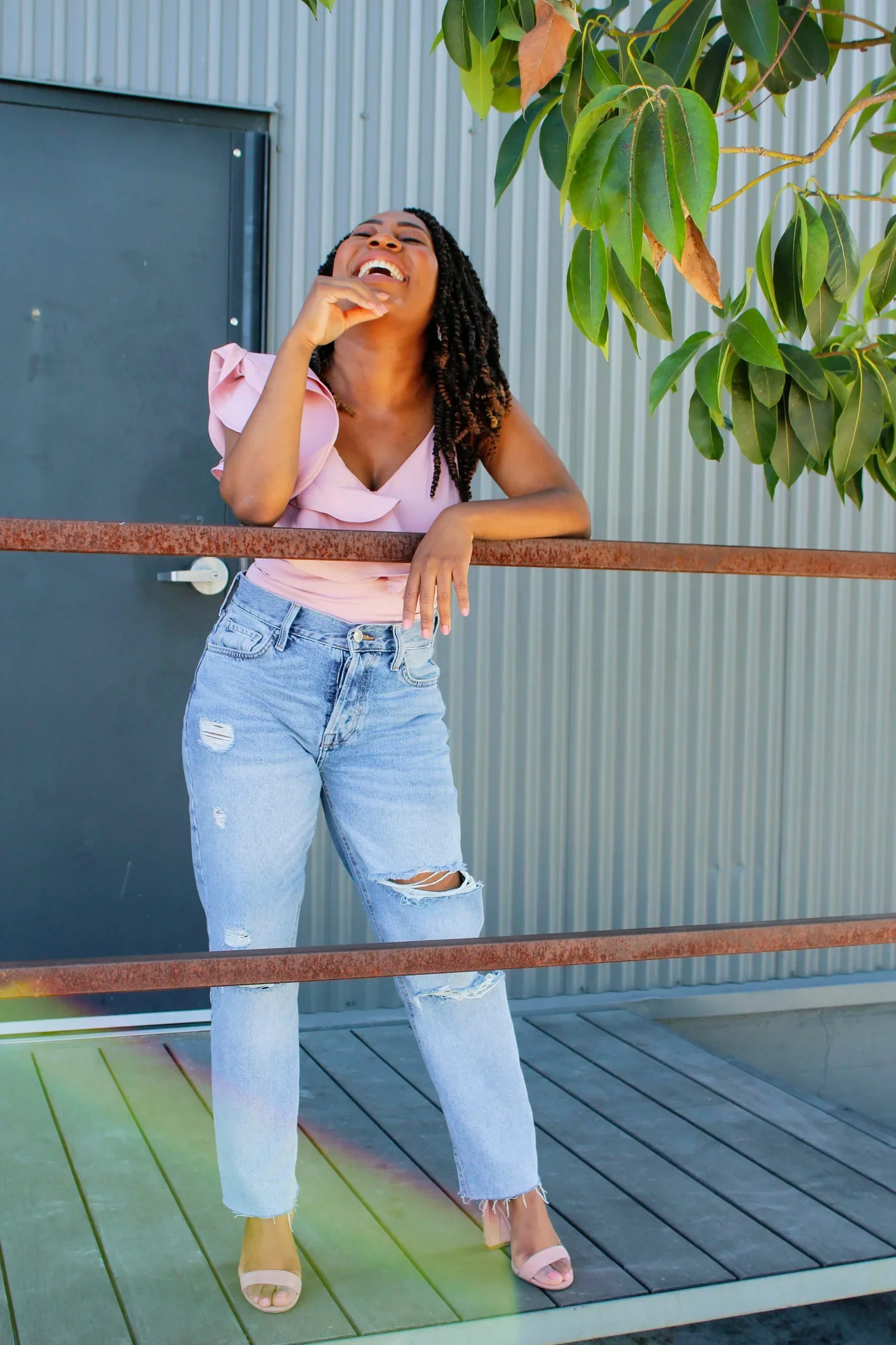A Black woman with dark curly hair laughing, wearing a pink blouse and ripped jeans, standing outdoors next to a green tree, rainbow, and a metal building, leaning on a rusted metal railing smiling towards the sky.