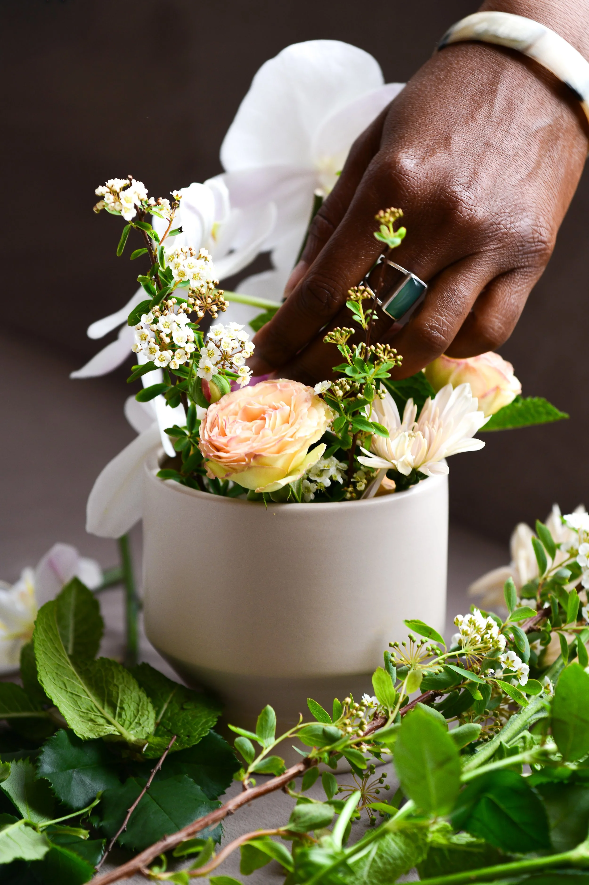 A person of color (POC) hand with a ring and bracelet arranging a bouquet of various flowers and greenery in a white vase.