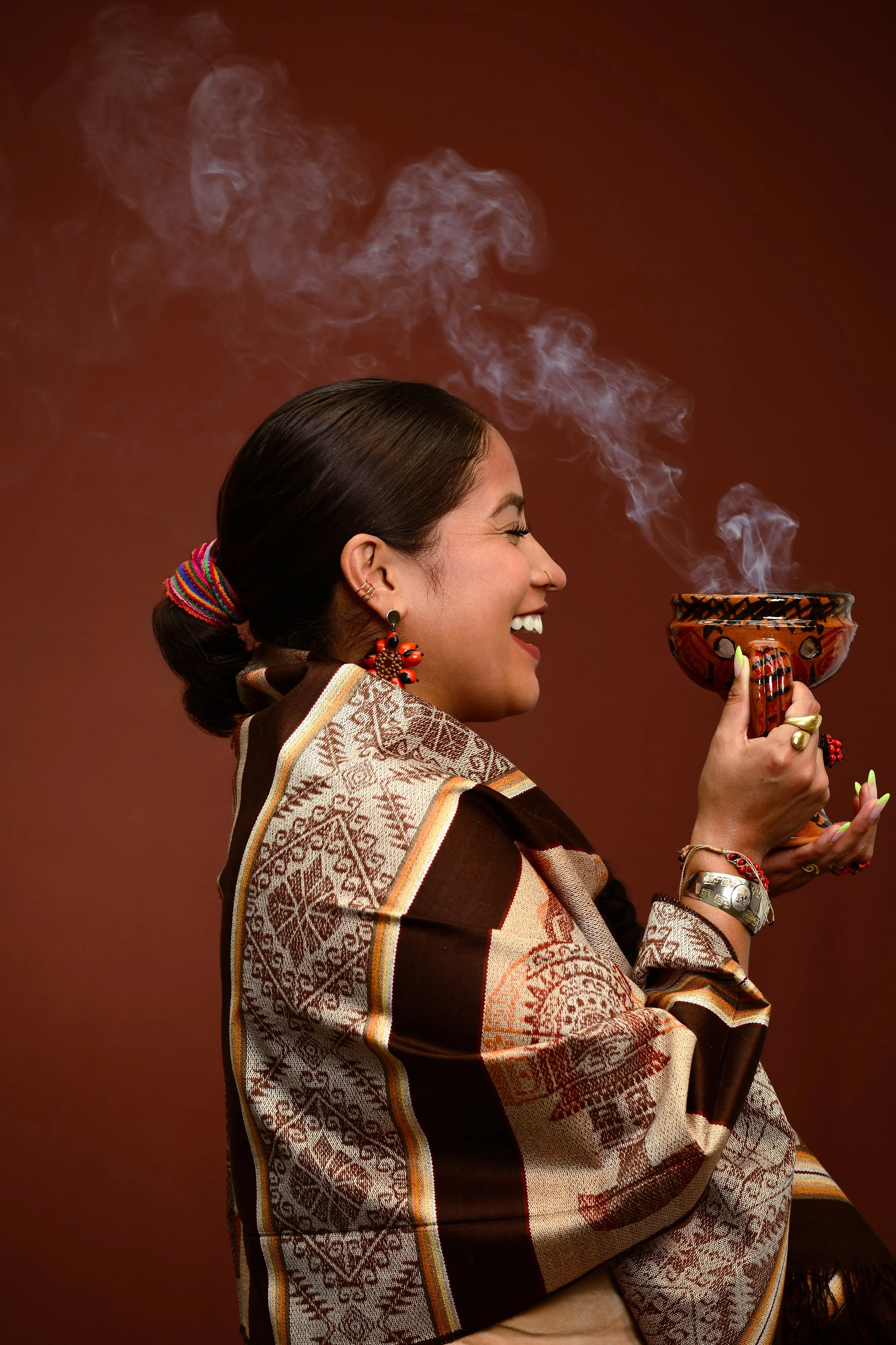 A woman with dark hair styled in a low bun, smiling and holding a decorated ceramic bowl with steam billowing from it. She is wearing earrings, bracelets, a ring, and a colorful scarf over her shoulder. The background is smoke or mist on a dark reddish-brown backdrop.