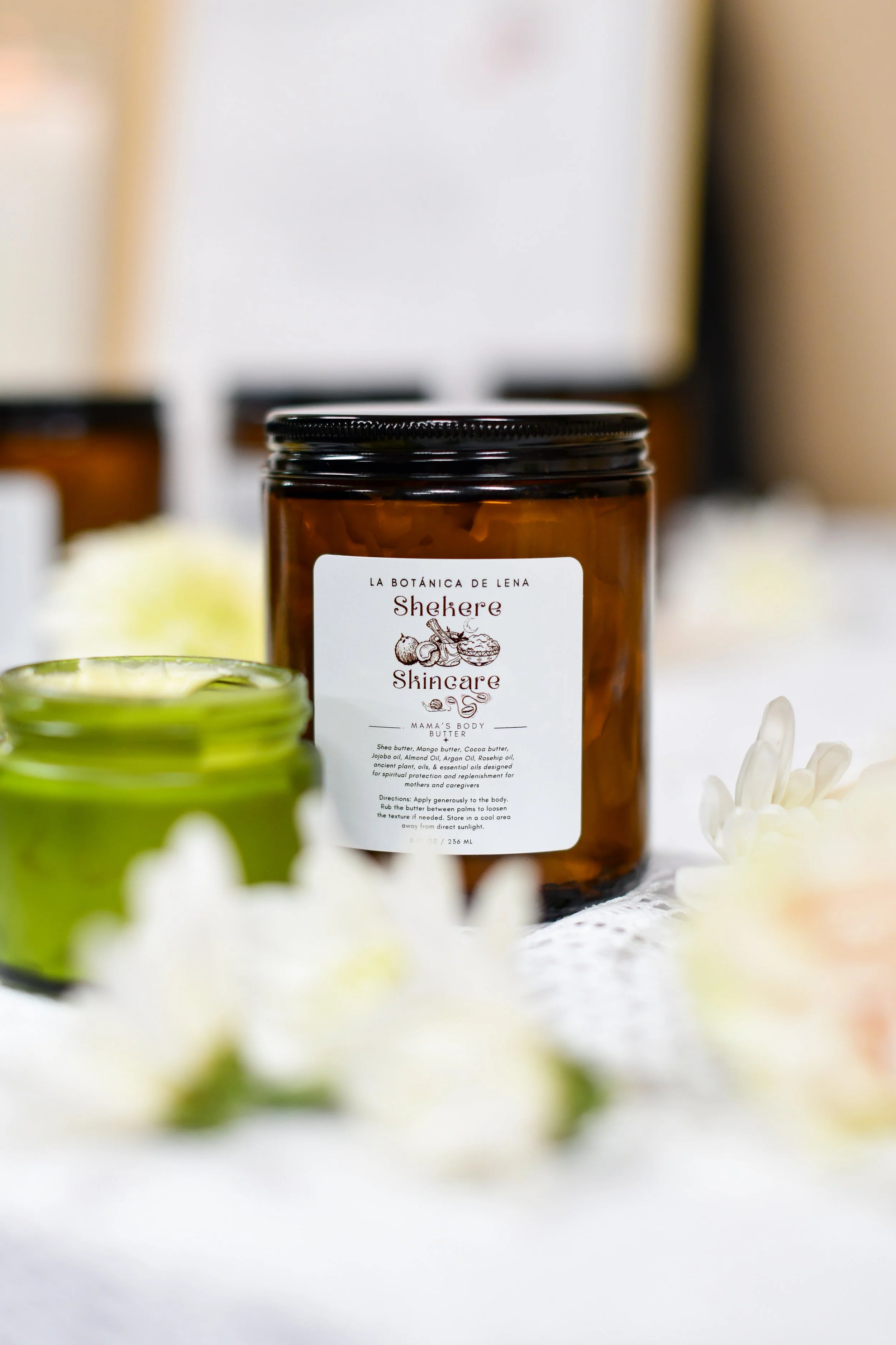 A close-up of a product brown glass jar labeled 'Shekere Skincare' with other skincare containers and white flowers surrounding it.