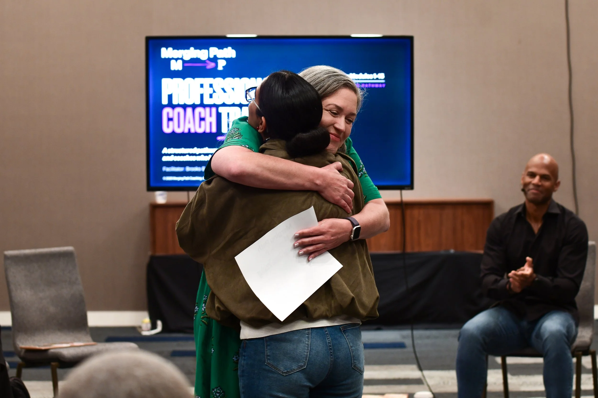 Two women hugging in a room with a large screen displaying a seminar or conference presentation. One woman has gray hair and the other has black hair, wearing glasses. Another person is seated to the side, clapping.
