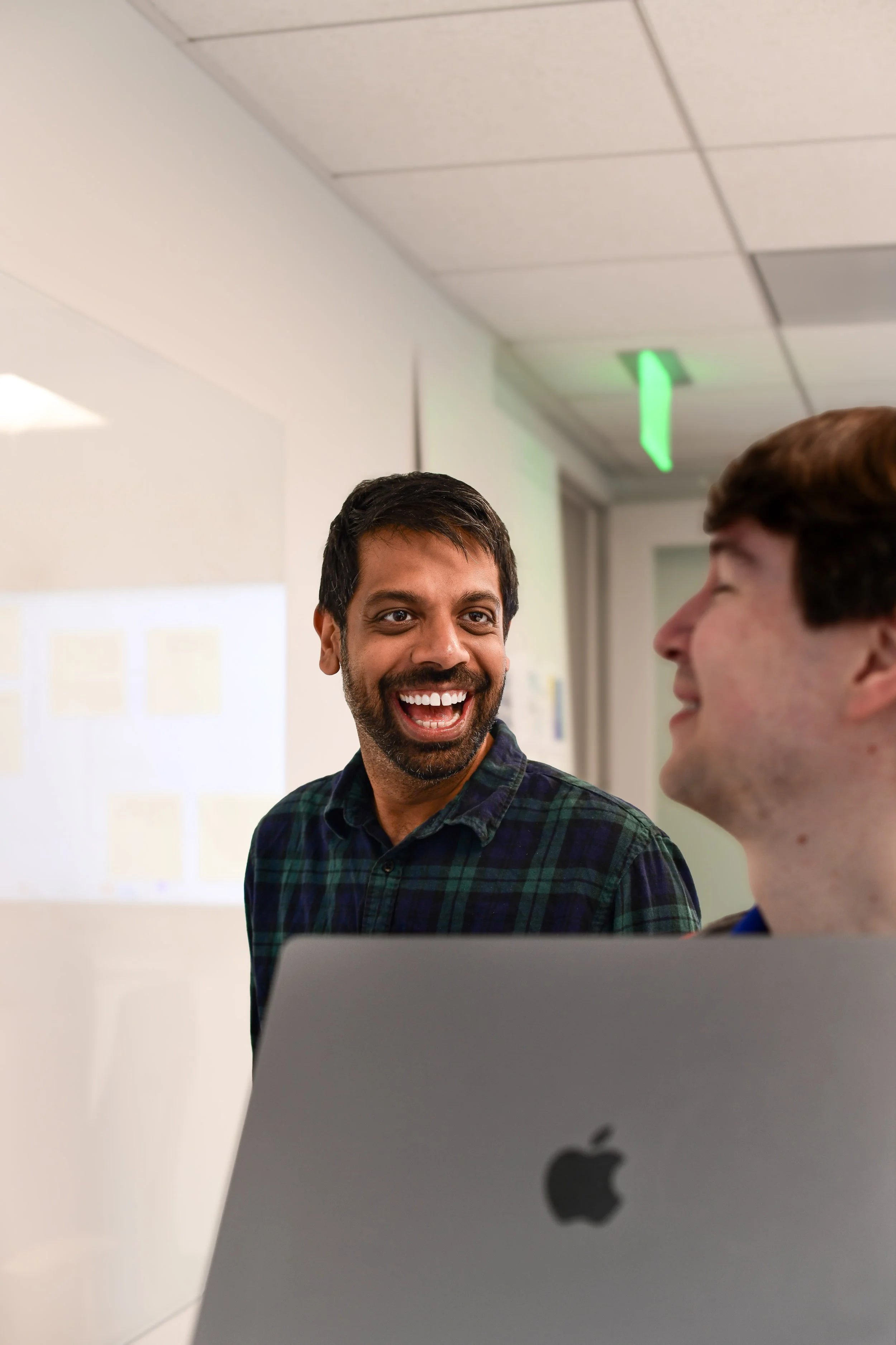 Two tech engineer men are having a conversation in an office space. One man is facing the camera with a big smile, while the other is turned to the side, listening. A laptop with an Apple logo is in the foreground.