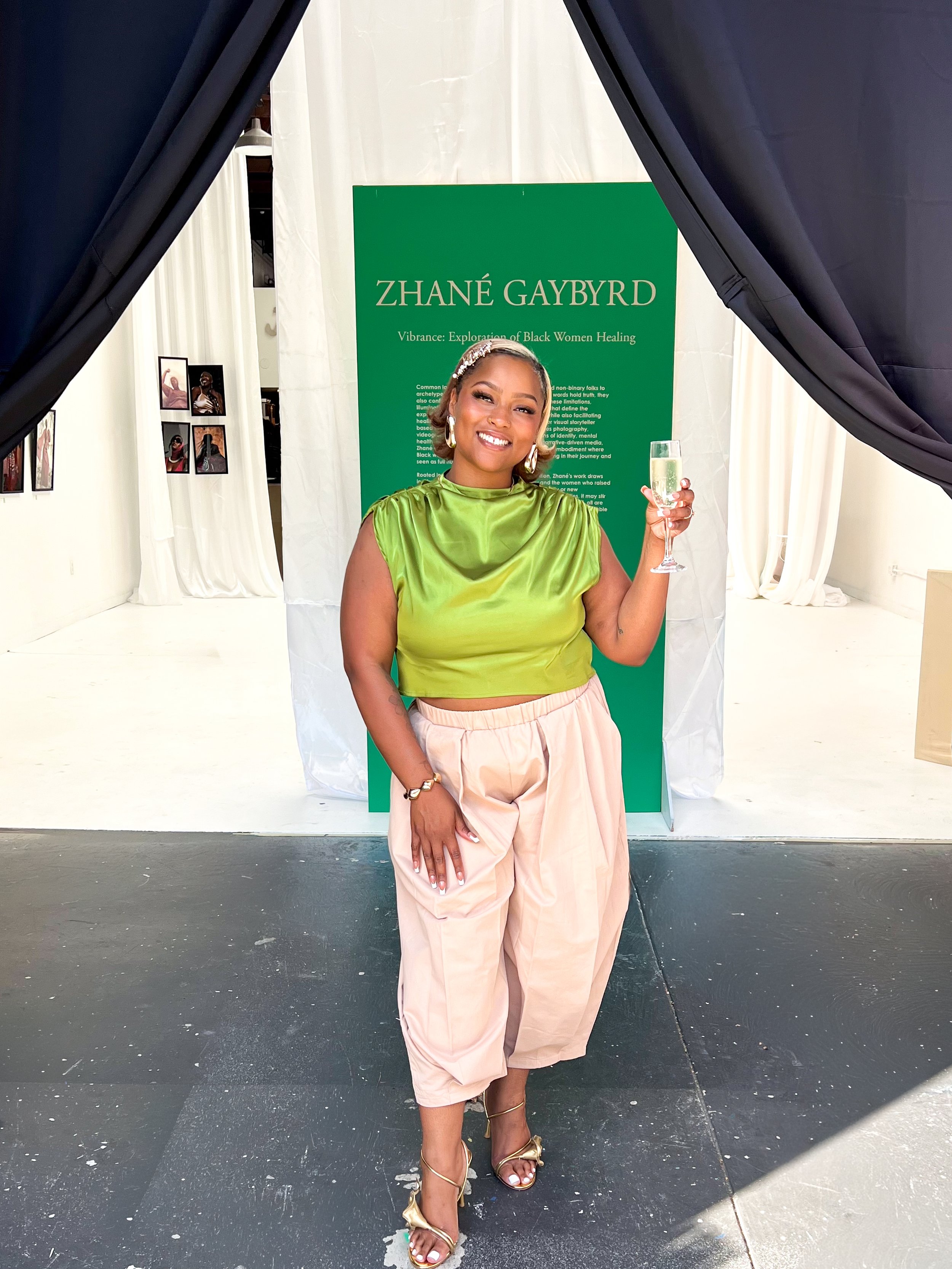 A woman standing in front of a green sign that reads 'Zhané Gaybyrd', holding a glass of champagne and smiling. She is dressed in a bright green satin top, beige pants, and gold high-heeled sandals, with large earrings and a hair accessory.