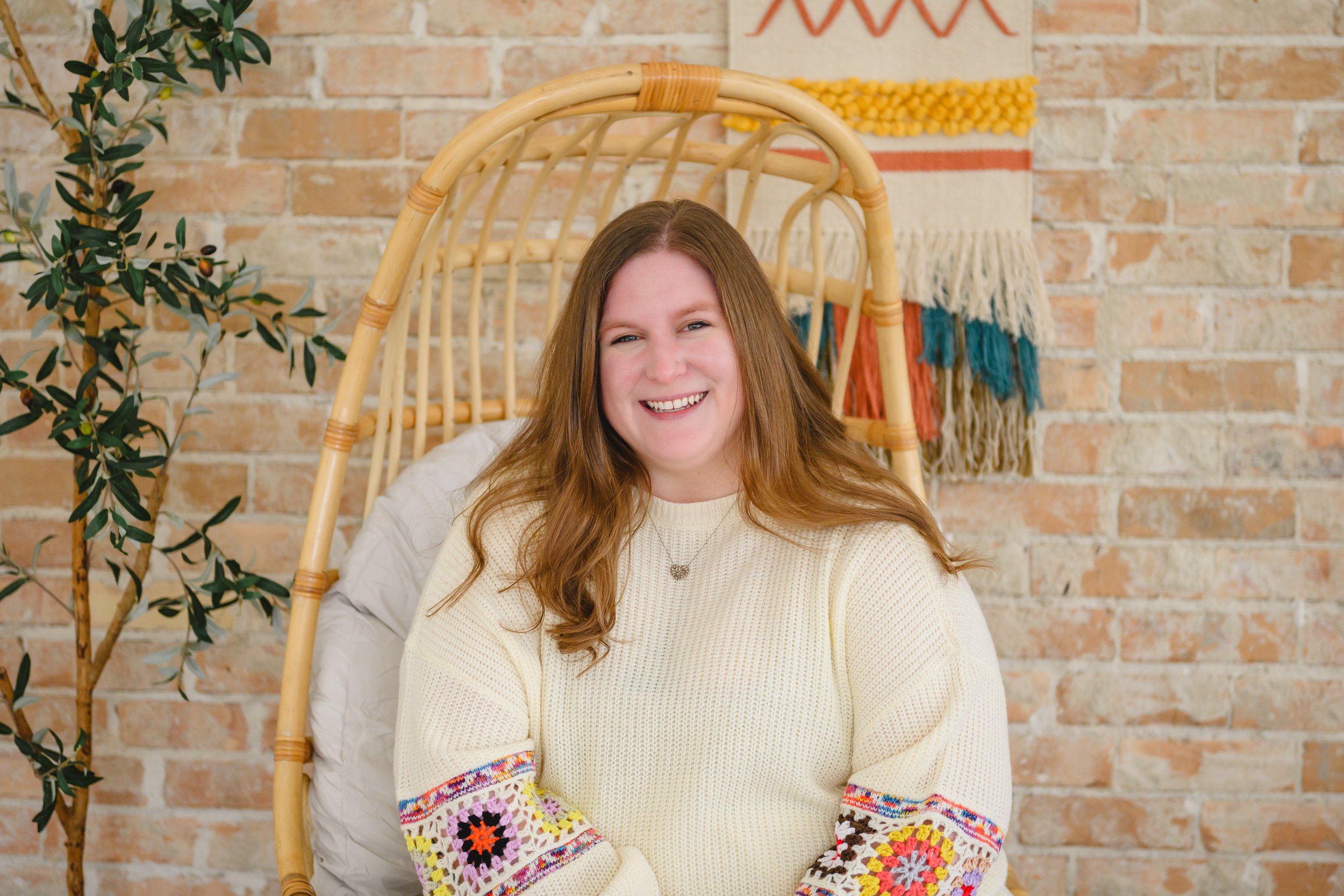 A woman with long red hair, smiling, sitting in a wicker chair with a white cushion, in front of a brick wall with a colorful woven wall hanging and a potted plant.