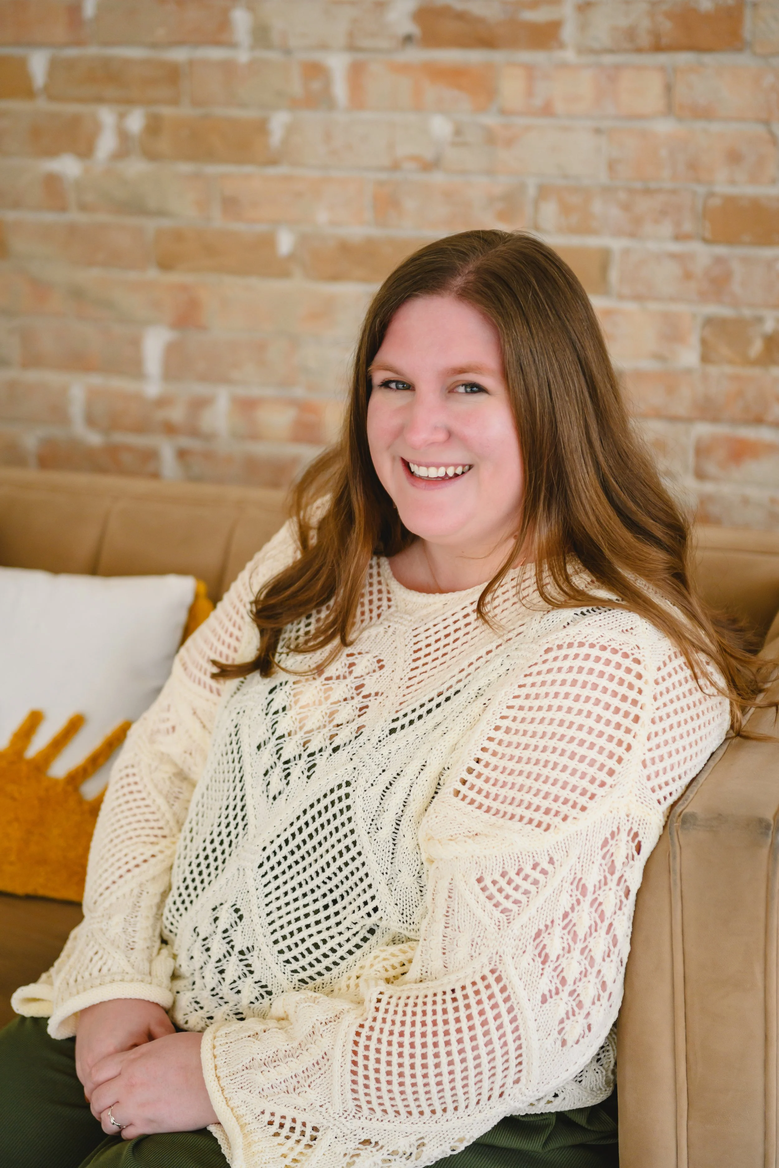 A woman with long, brown hair smiling while sitting on a beige couch in front of a brick wall, wearing a cream knit sweater.