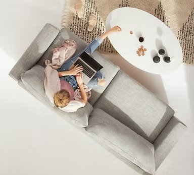 A person sitting on a gray sofa playing on a laptop, with snacks and drinks on a white coffee table nearby.