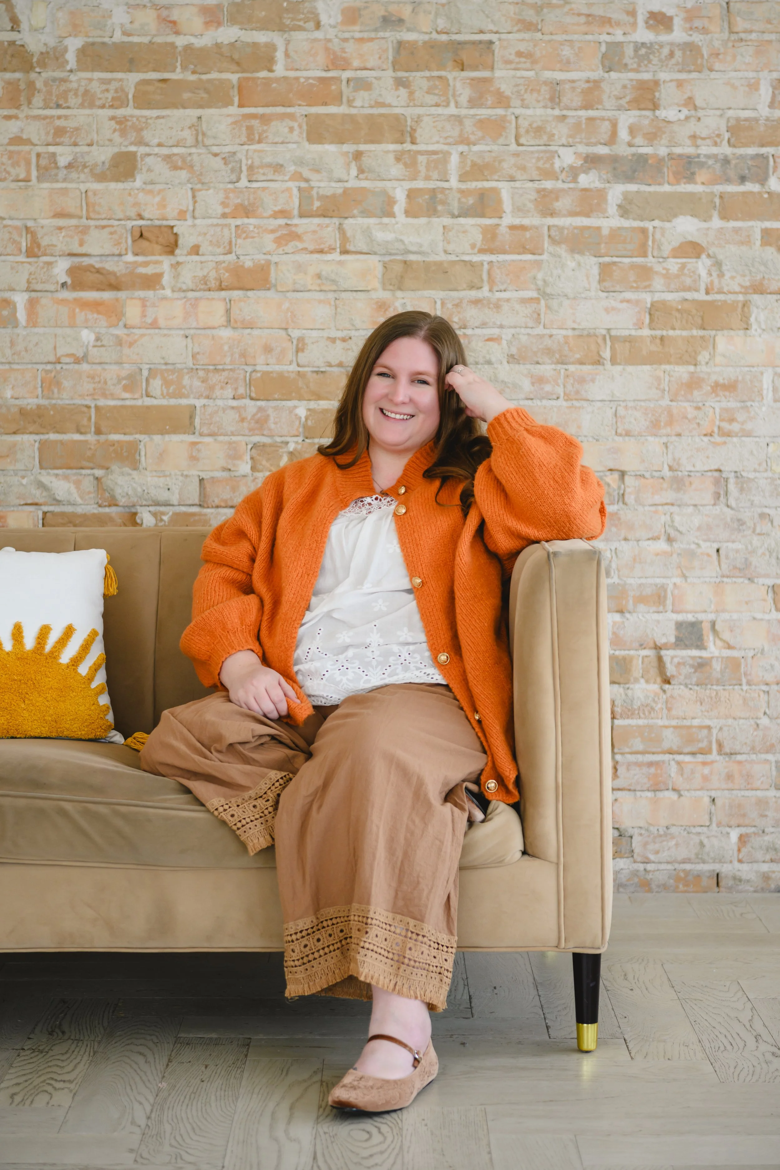 A woman sitting on a beige sofa against a brick wall, smiling while wearing an orange cardigan, white blouse, beige skirt, and ballet flats.