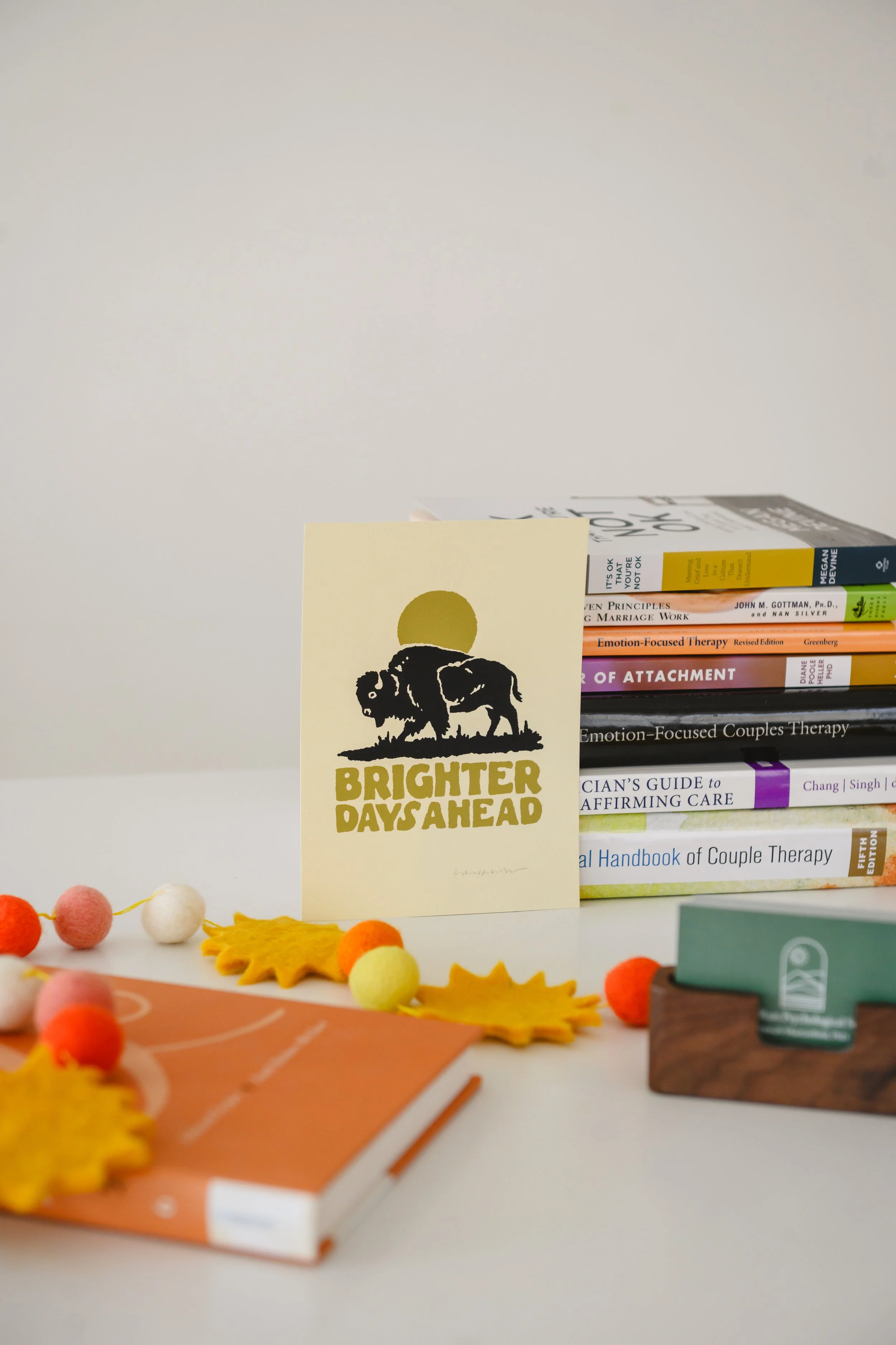 Books stacked on a white surface, a card with a bison and sun illustration that reads 'Brighter Days Ahead', colorful felt leaf and pom-pom decorations, and a small tray with business cards.
