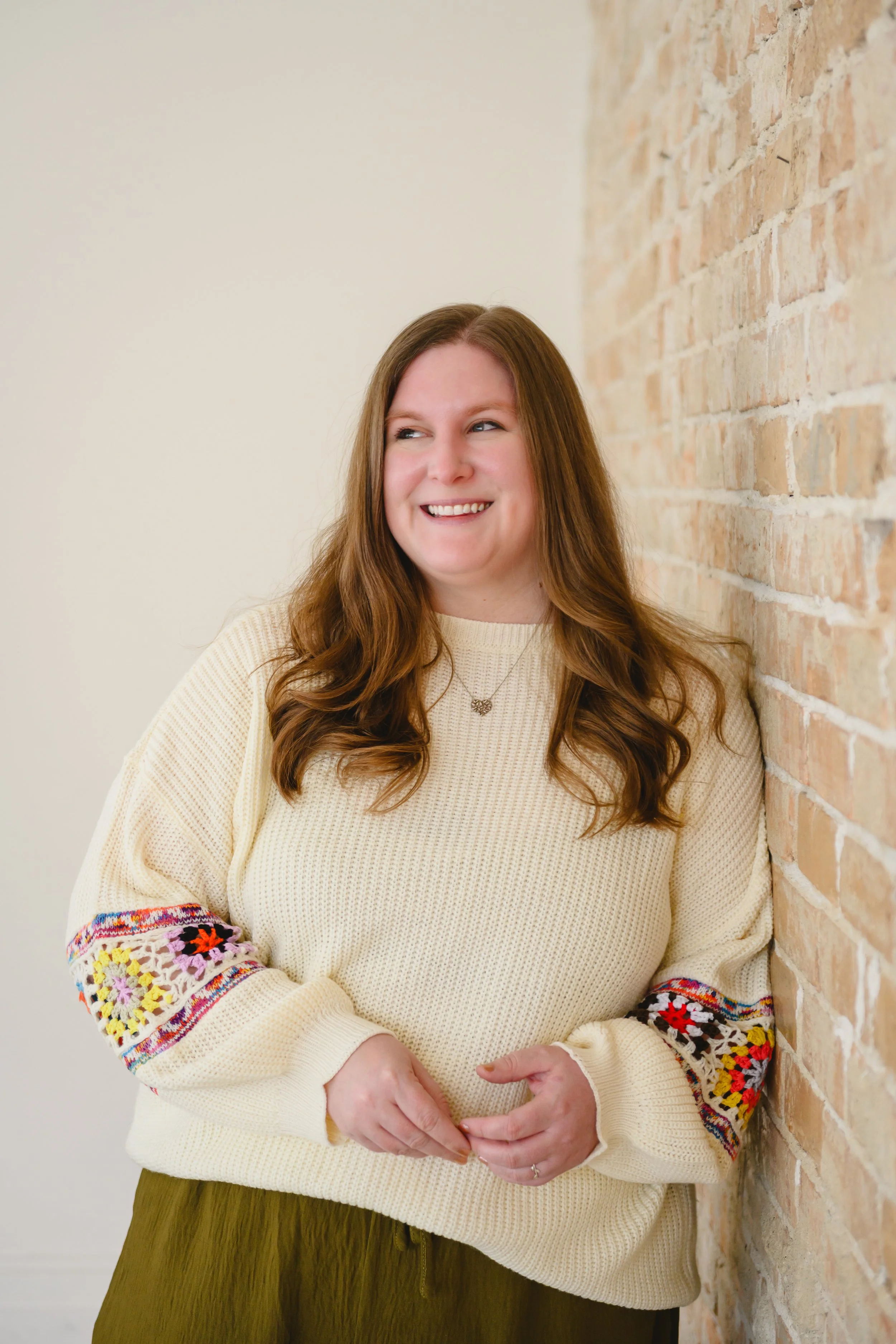 A woman with long, wavy red hair smiling and leaning against a brick wall, wearing a cream sweater with embroidered colorful patterns on the sleeves and an olive green skirt.