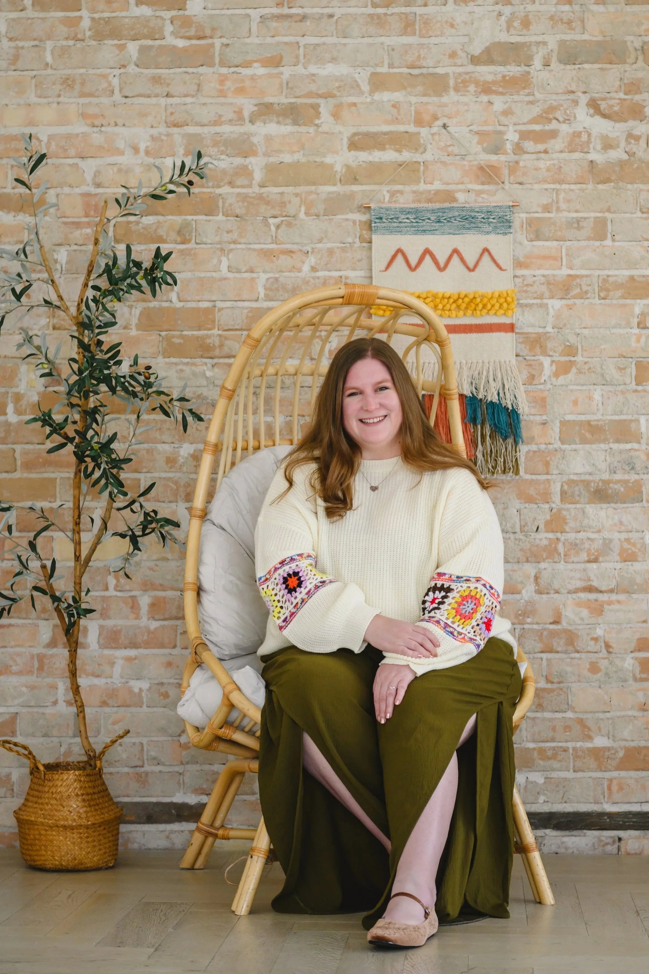 A woman sitting on a rattan chair in front of a brick wall, smiling. There is a potted plant to her left and a woven wall hanging behind her.