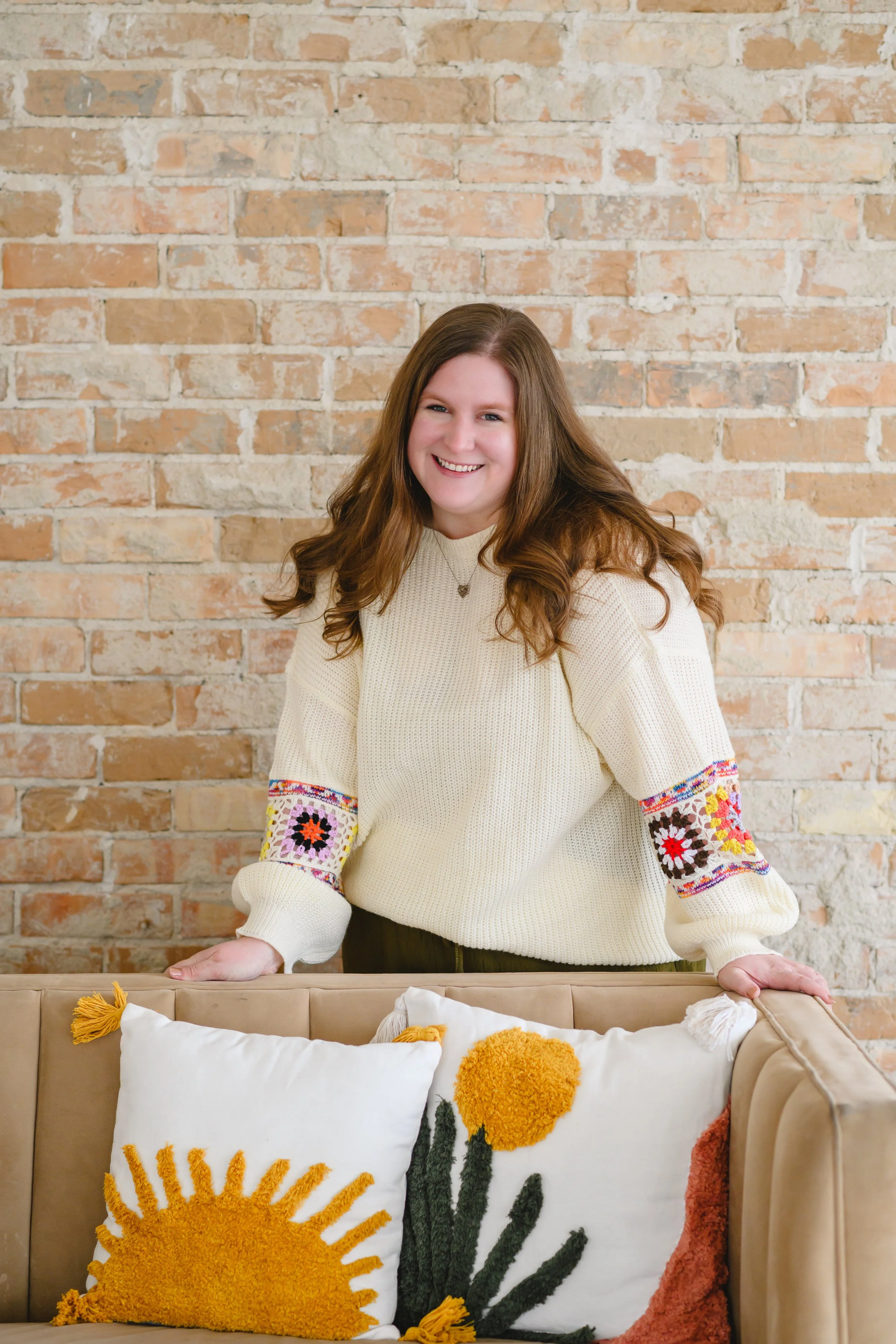 A smiling woman with long, wavy brown hair stands behind a beige sofa decorated with two pillows featuring sun and flower designs. She is wearing a cream sweater with colorful embroidery on the sleeves and a small necklace, with a brick wall in the background.