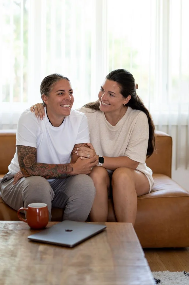 Two women sitting on a couch, smiling and holding hands, in a bright living room with large windows, a coffee table with a laptop and a mug in front of them.