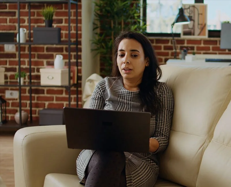 A woman sitting on a cream-colored sofa using a laptop in a modern living room with brick walls, green plants, and shelves with decor items.