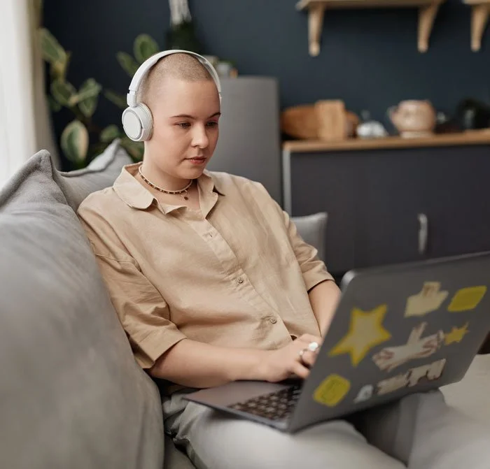 A young person with a shaved head wearing headphones, sitting on a gray couch, using a laptop in a cozy indoor setting.