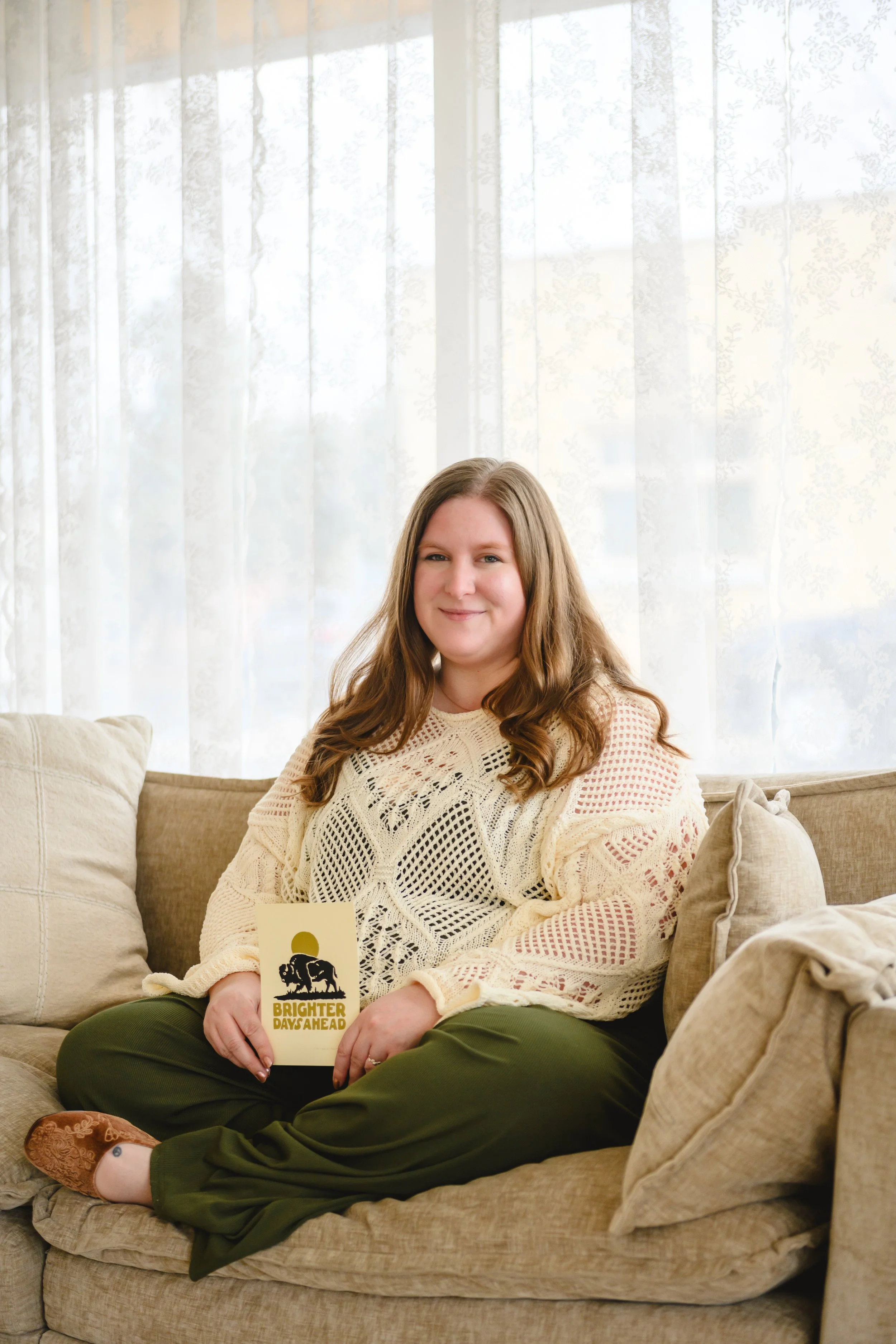 A woman with long, wavy brown hair sitting on a beige sofa in front of a window with sheer white curtains, holding a small sign that reads 'Brighter Days Ahead' with a dog silhouette and a sun design.
